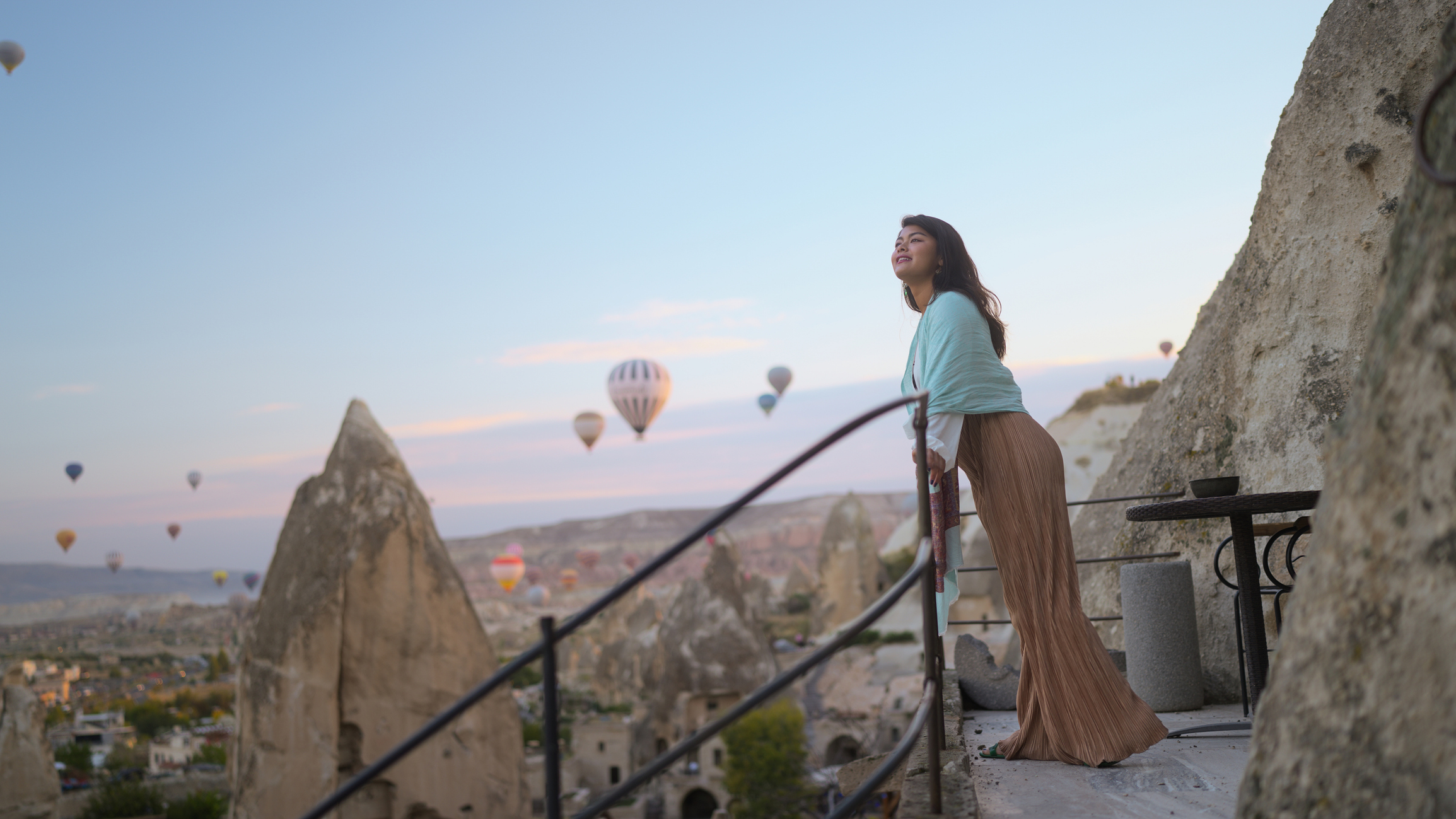 A woman stands on a balcony overlooking a valley with hot air balloons in the sky at sunset.