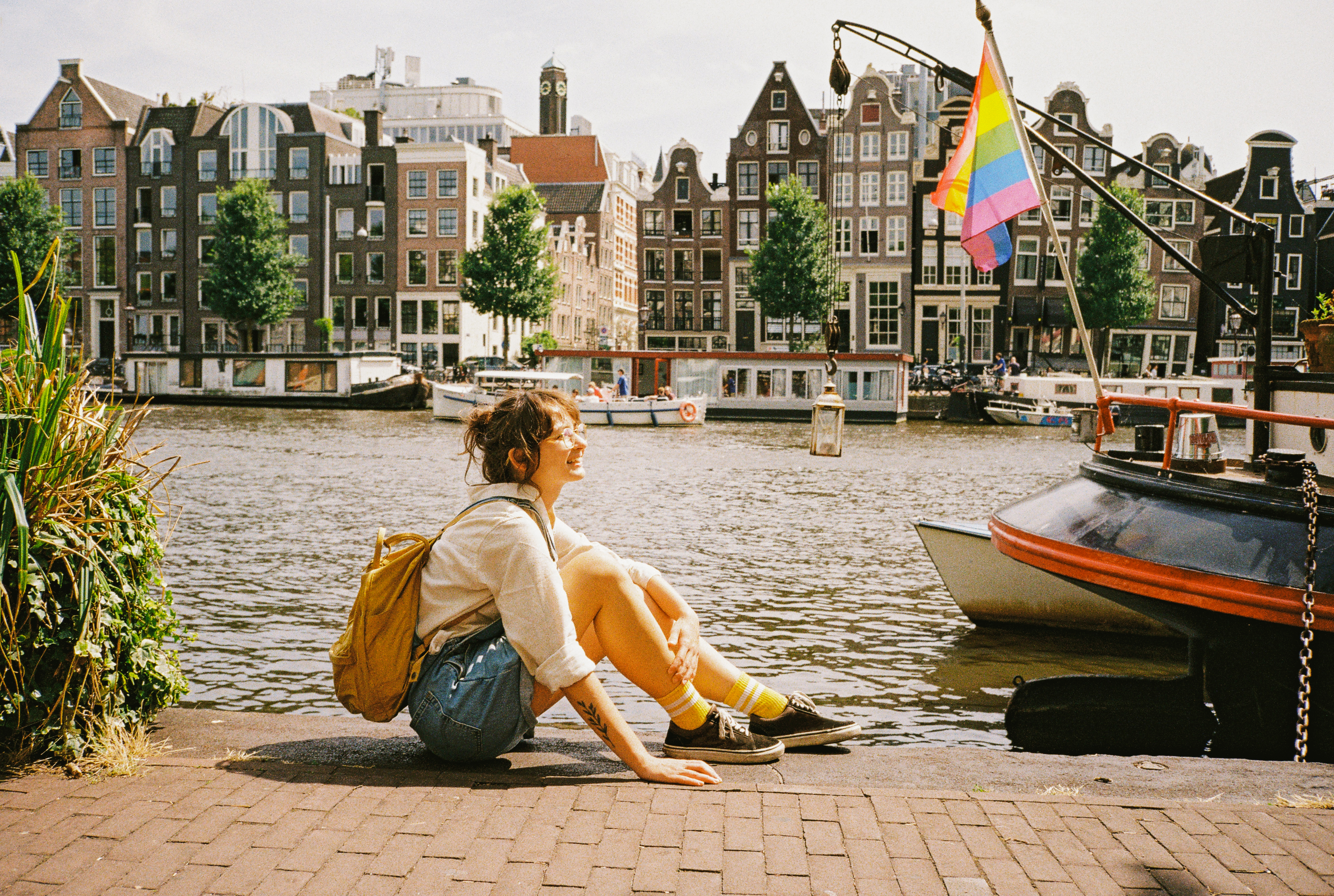 Person sitting by a canal in Amsterdam with colorful buildings and a rainbow flag in the background on a sunny day.