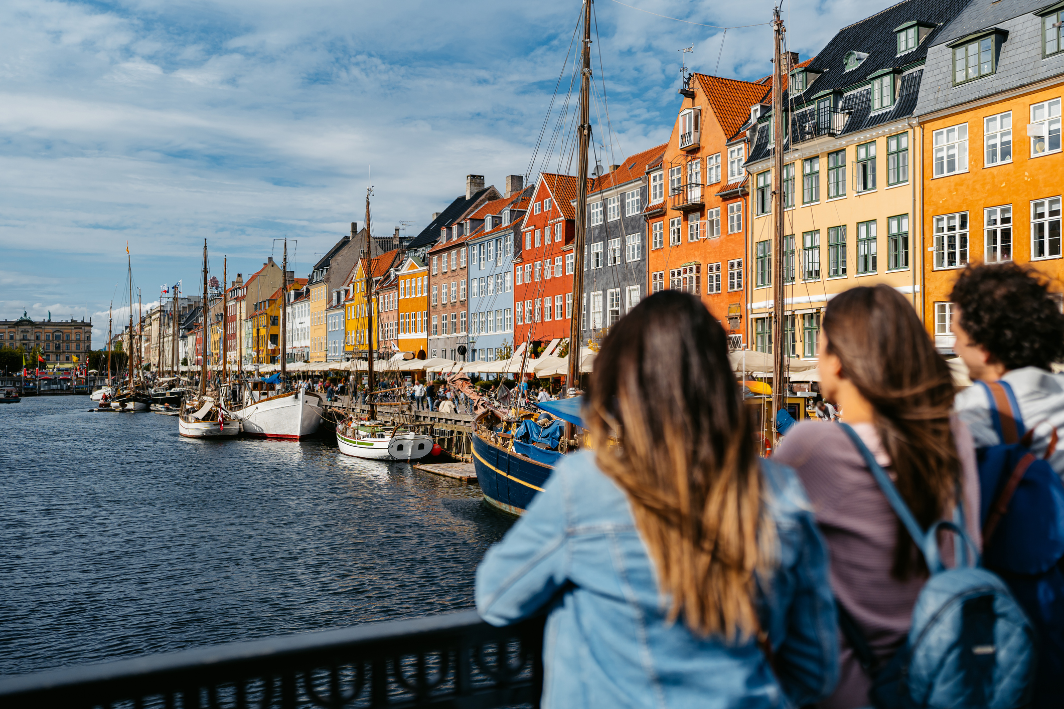 Three people stand by a canal, gazing at colorful buildings and boats in a scenic waterfront view.