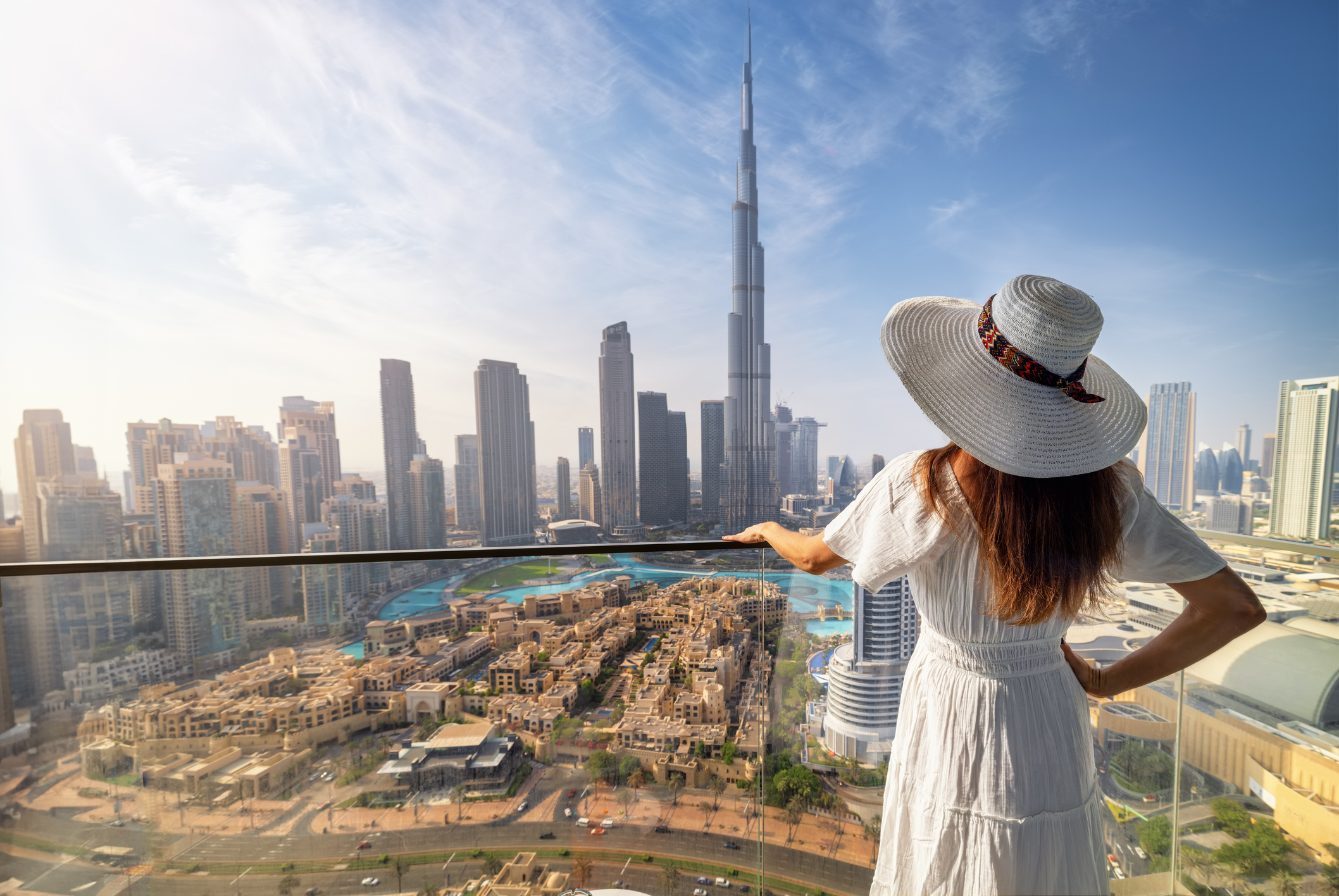 A woman in a white dress and hat gazes at a city skyline featuring the Burj Khalifa in the background.