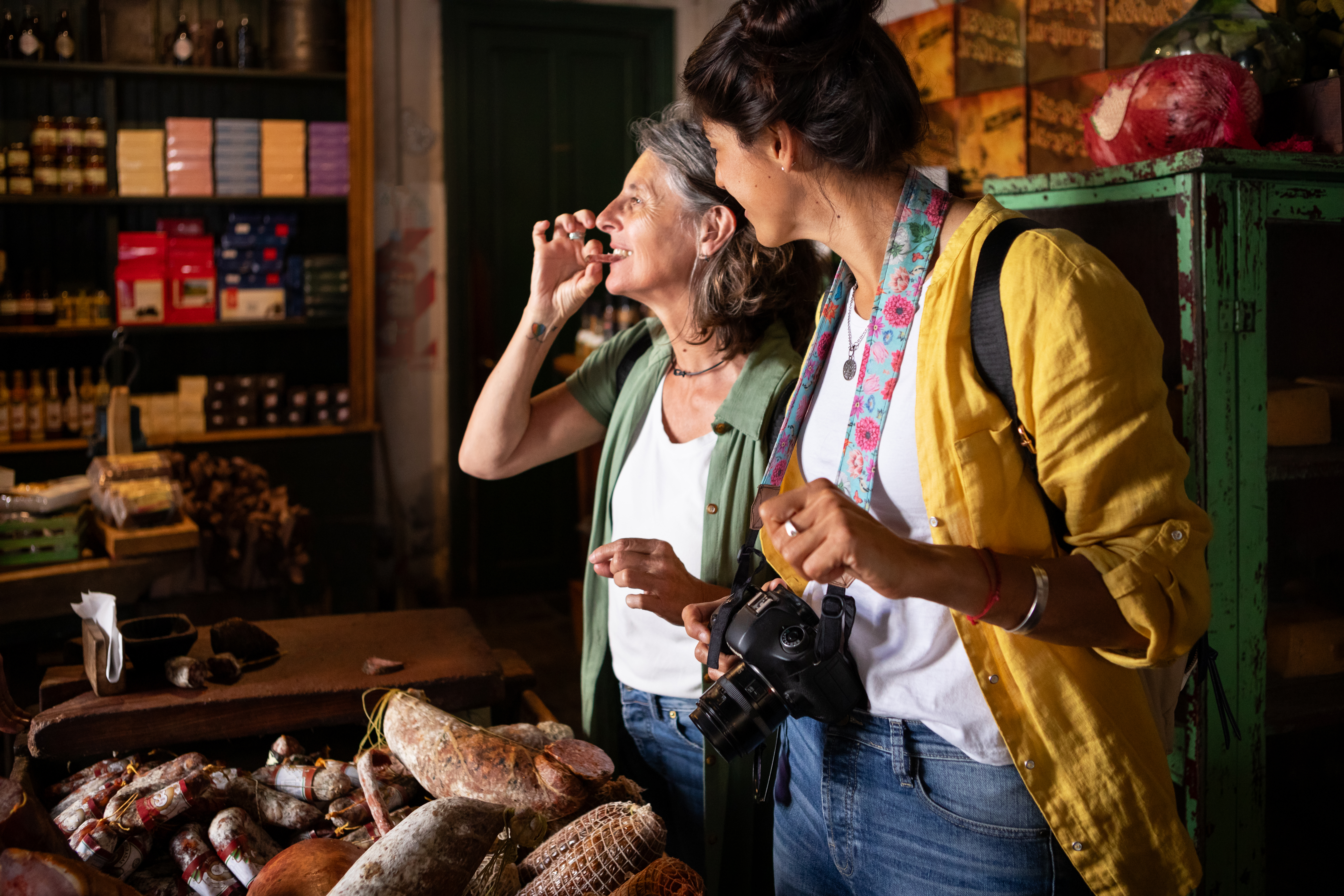 Two women enjoy a moment in a market, surrounded by various cured meats and goods, one holding a camera.