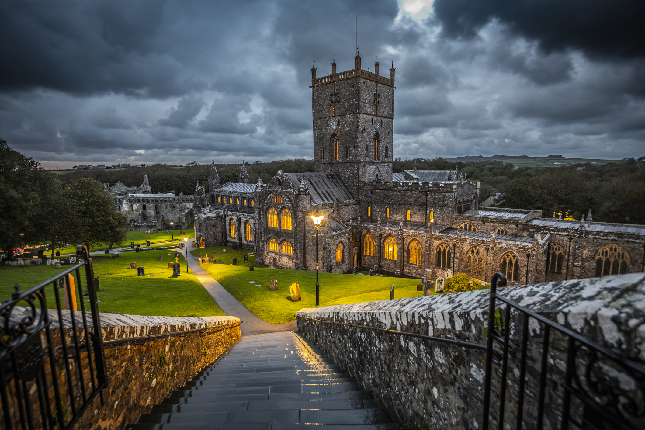 Historic stone church with a tall tower, surrounded by trees and glowing lights, under a dramatic cloudy sky.
