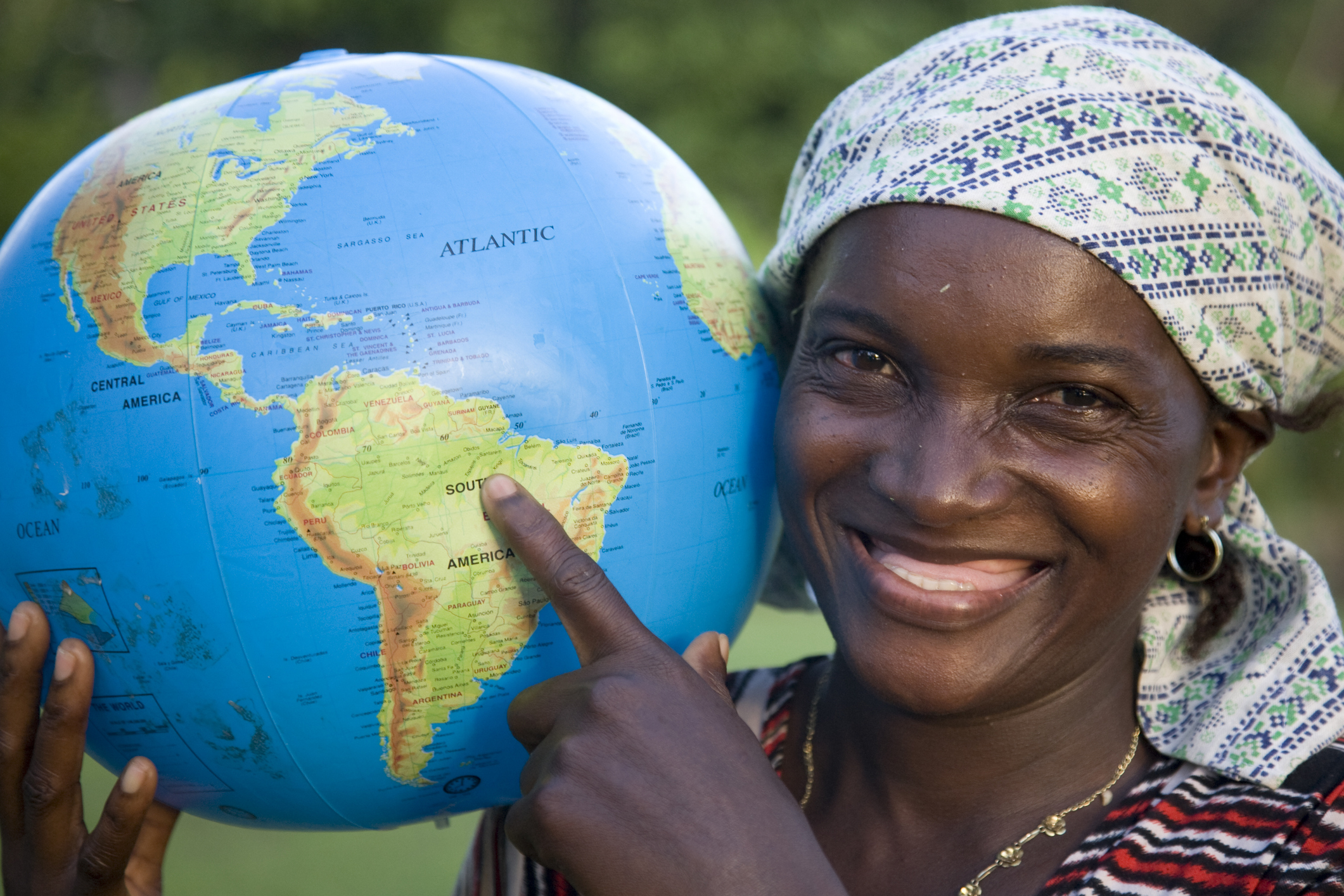 Smiling woman pointing at a globe, highlighting South America, against a natural backdrop.