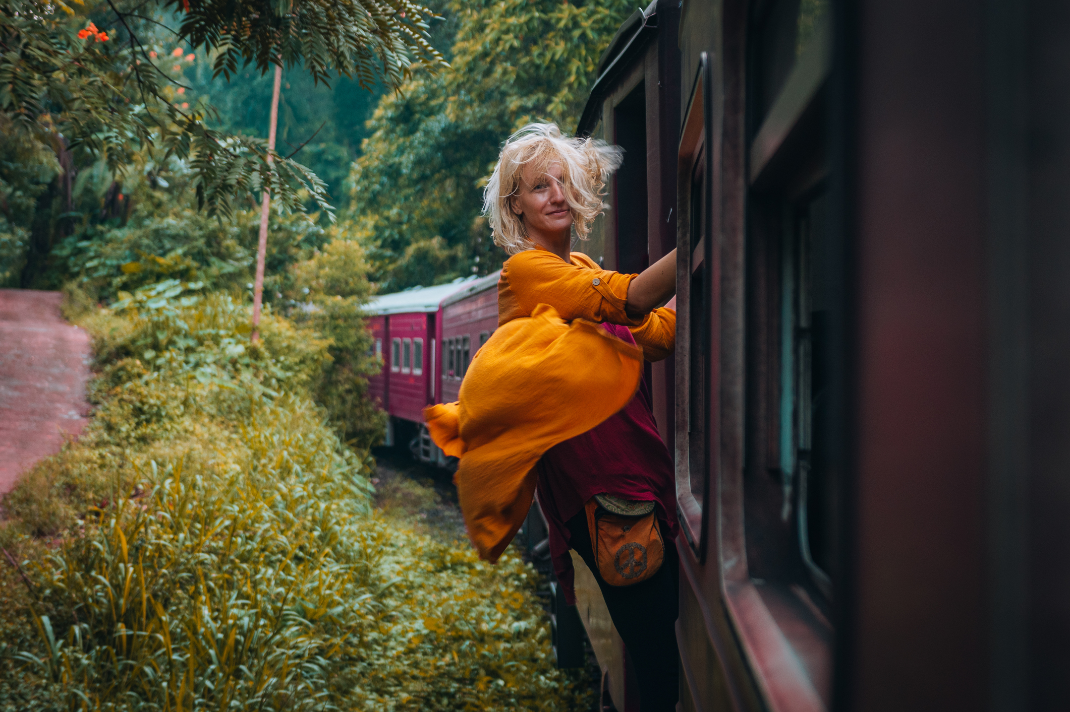 A person in a bright yellow dress leans out of a train window, surrounded by lush greenery and a second train in the background.