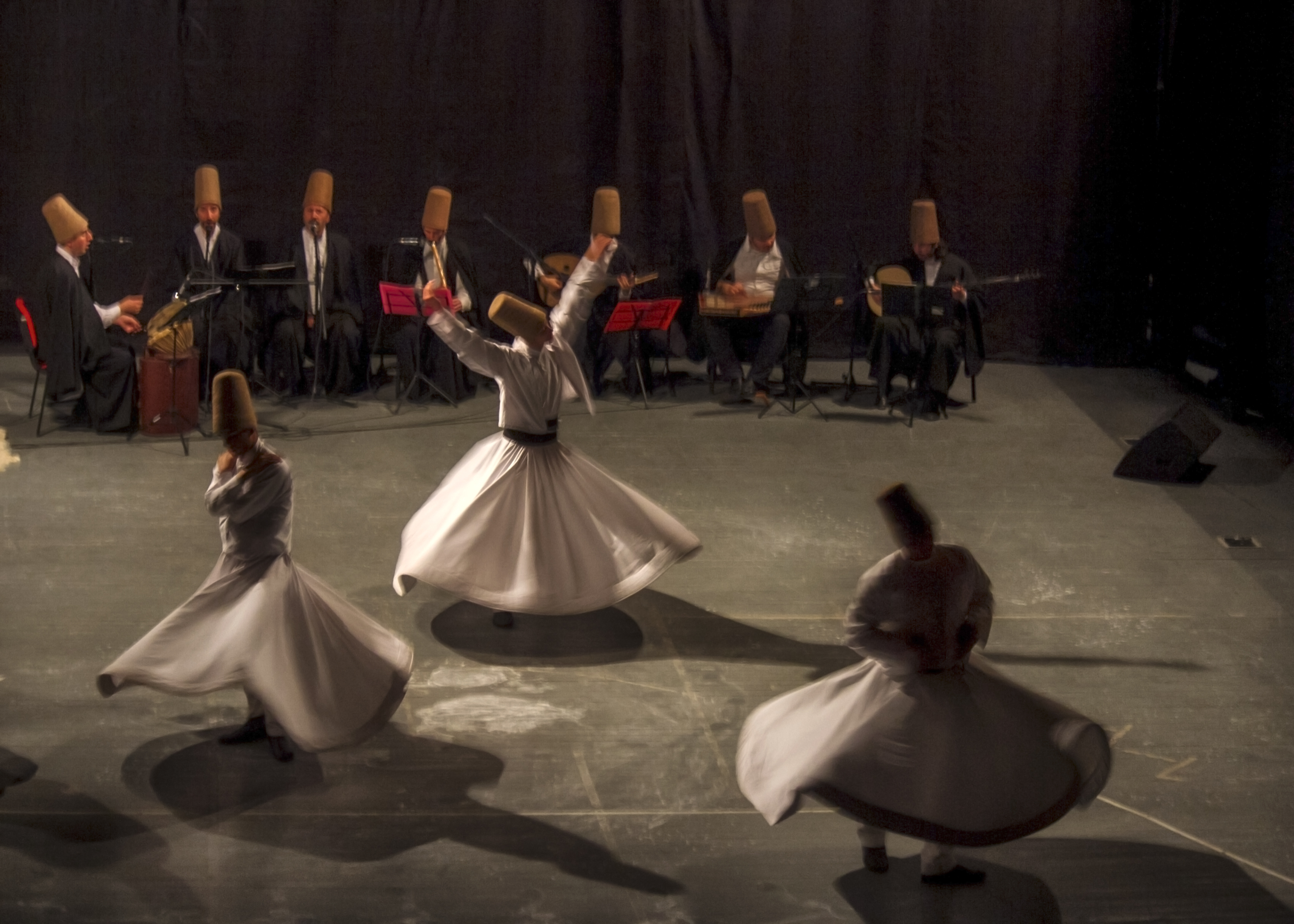 Whirling dervishes perform in front of musicians, all wearing traditional hats, on a dimly lit stage.