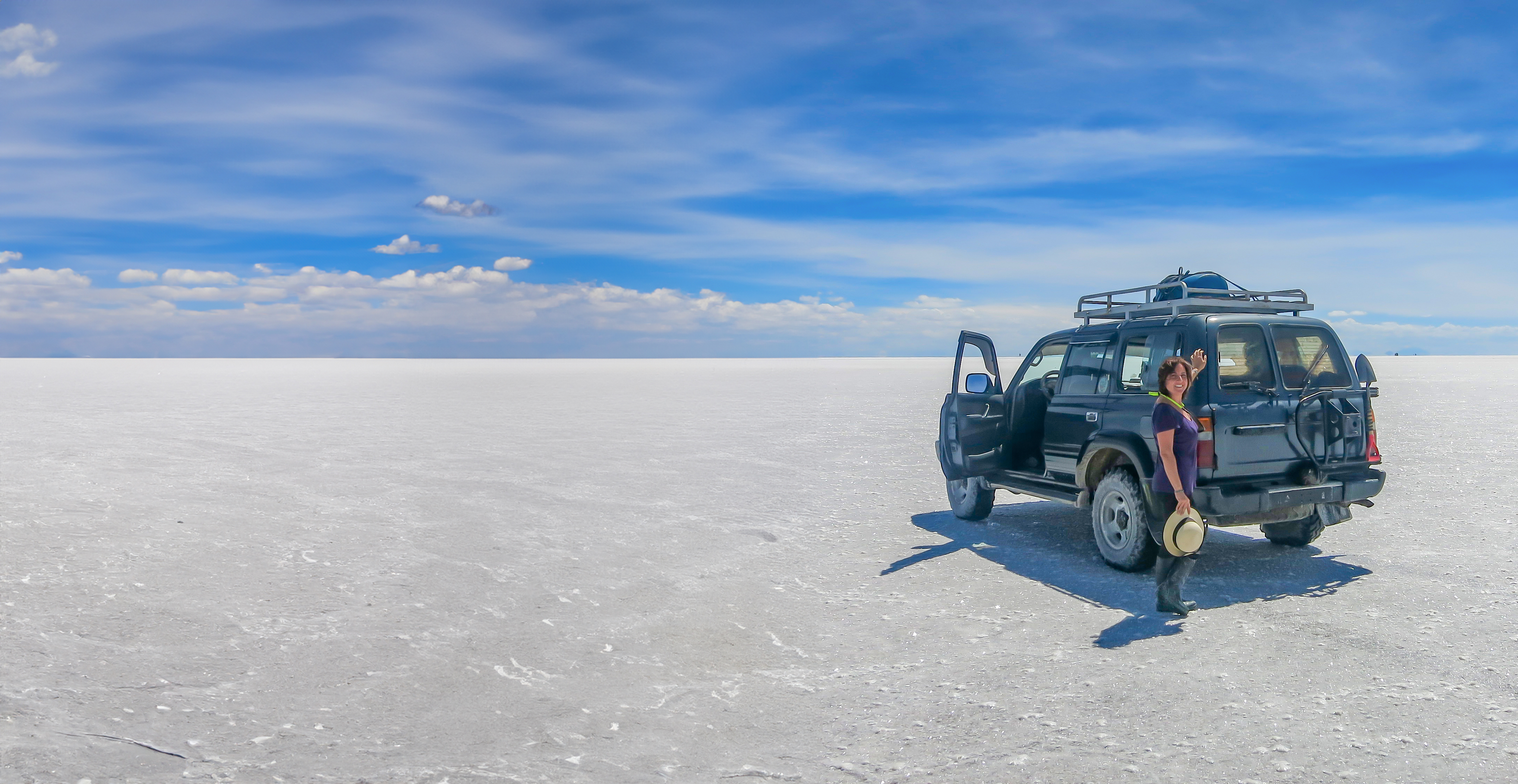 A person stands next to a parked SUV on a vast white salt flat under a blue sky with scattered clouds.