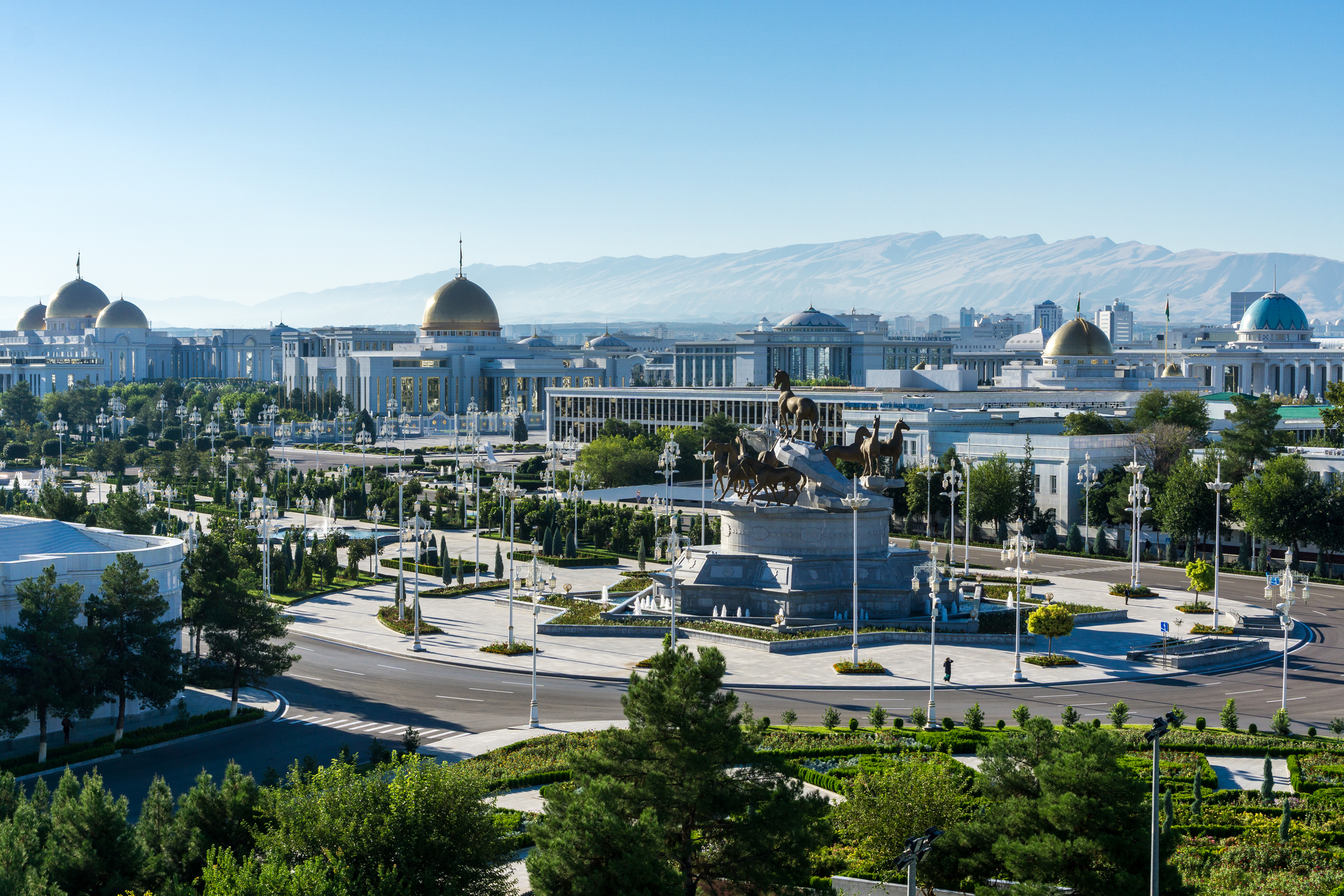 Panoramic view of a city with modern architecture, a large monument, and mountains in the background under clear blue skies.