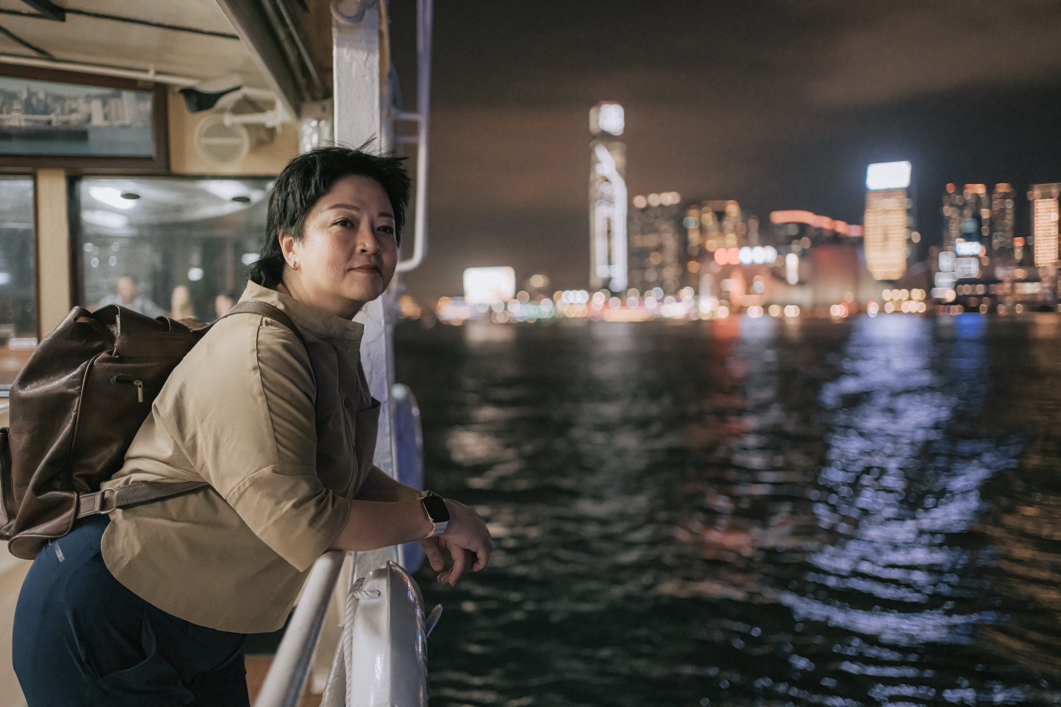 A person leans on a railing of a boat, gazing at a city skyline illuminated at night. Water ripples below.