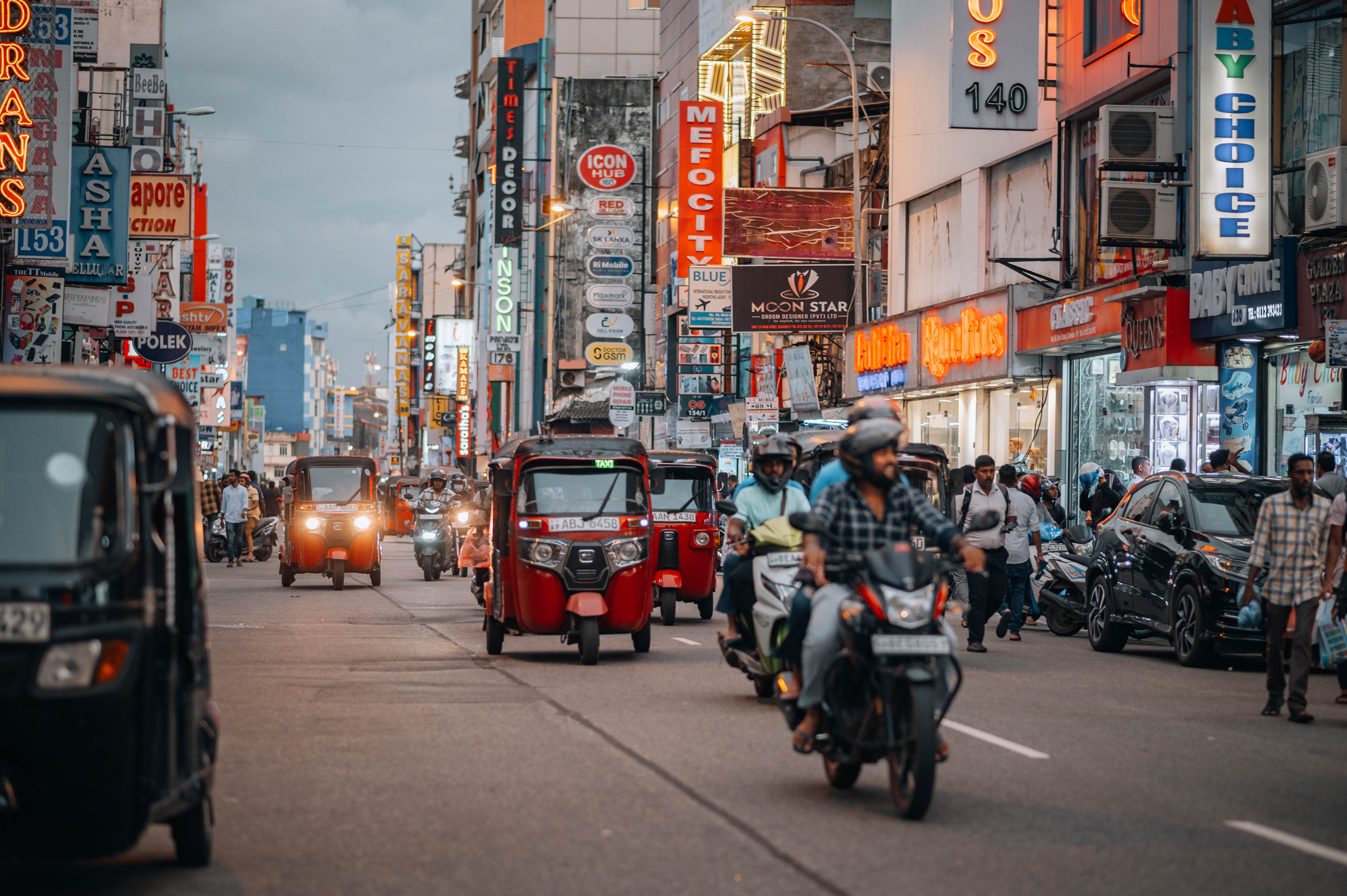 Busy street scene with tuk-tuks, motorcycles, and colorful storefronts illuminated by neon signs.