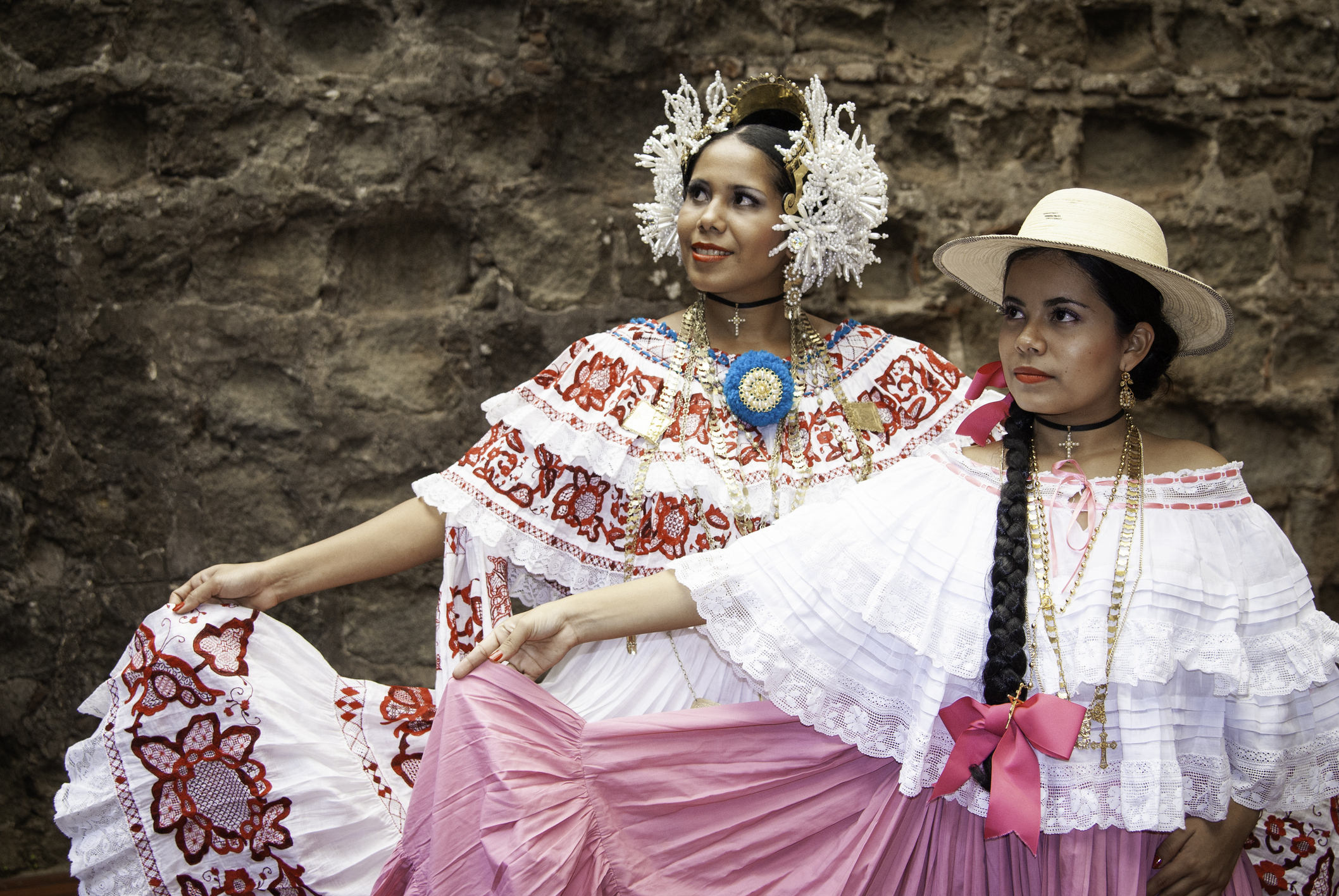 Two women in traditional Mexican dresses pose together, showcasing vibrant colors and cultural accessories against a stone backdrop.