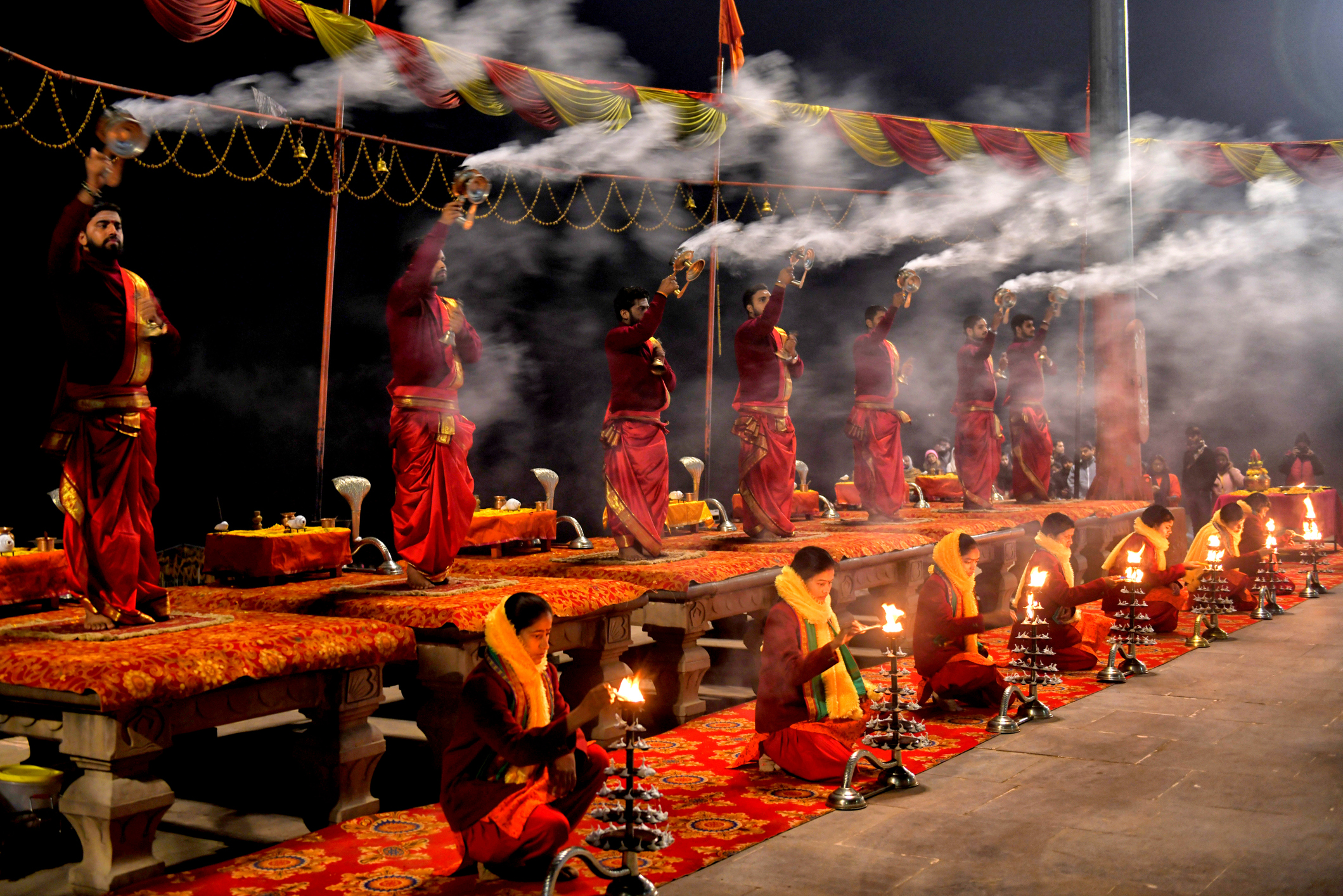 A group of individuals in traditional attire perform a ritual with lamps and incense, surrounded by smoke and bright decorations.