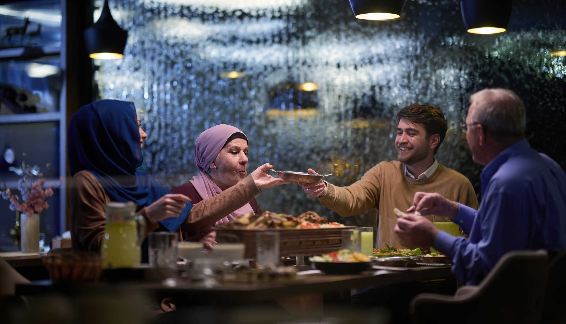 A group of four people dining together, sharing food, with a cozy atmosphere and a rain-streaked window in the background.