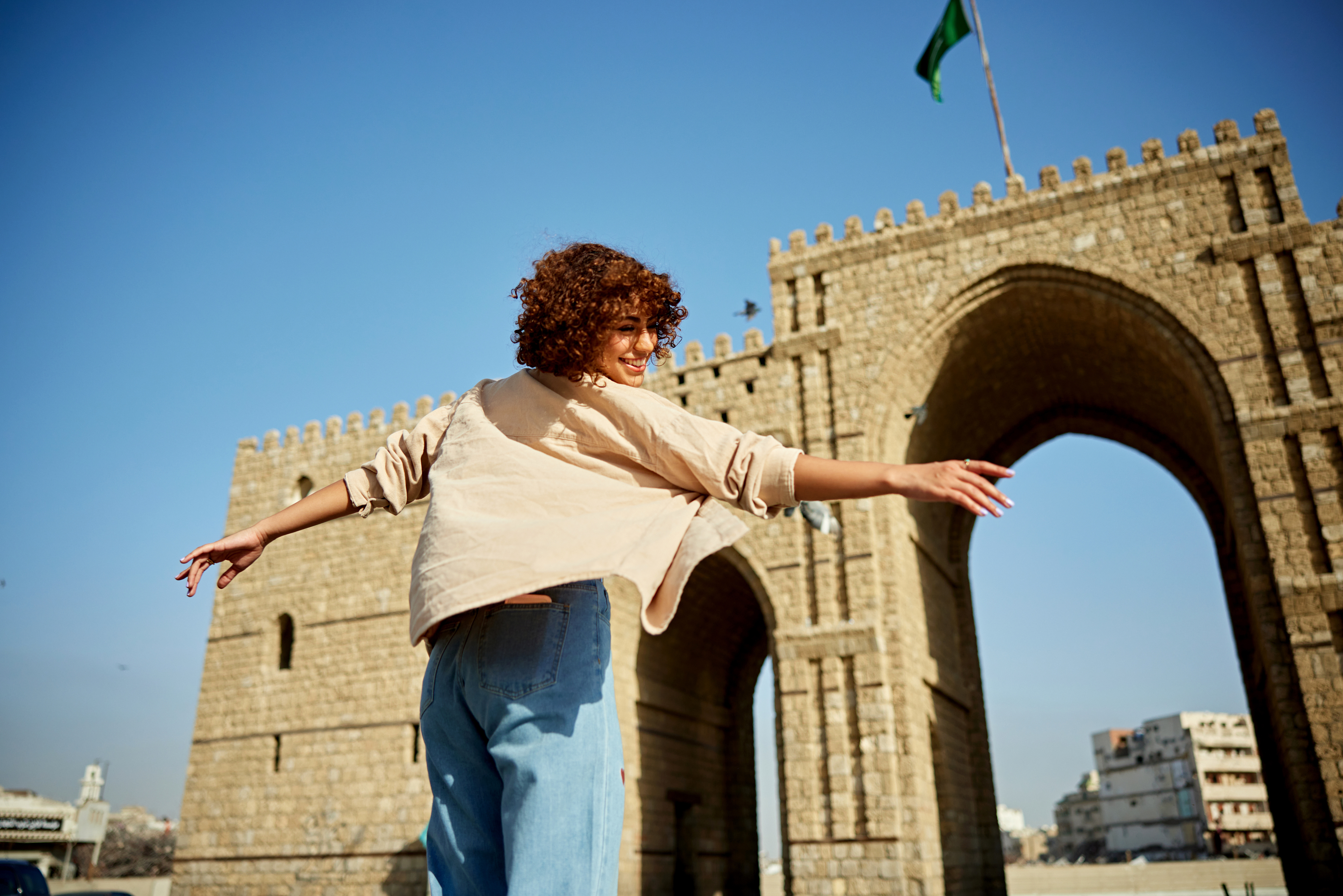 A person with curly hair joyfully spins in front of a large stone archway against a clear blue sky.