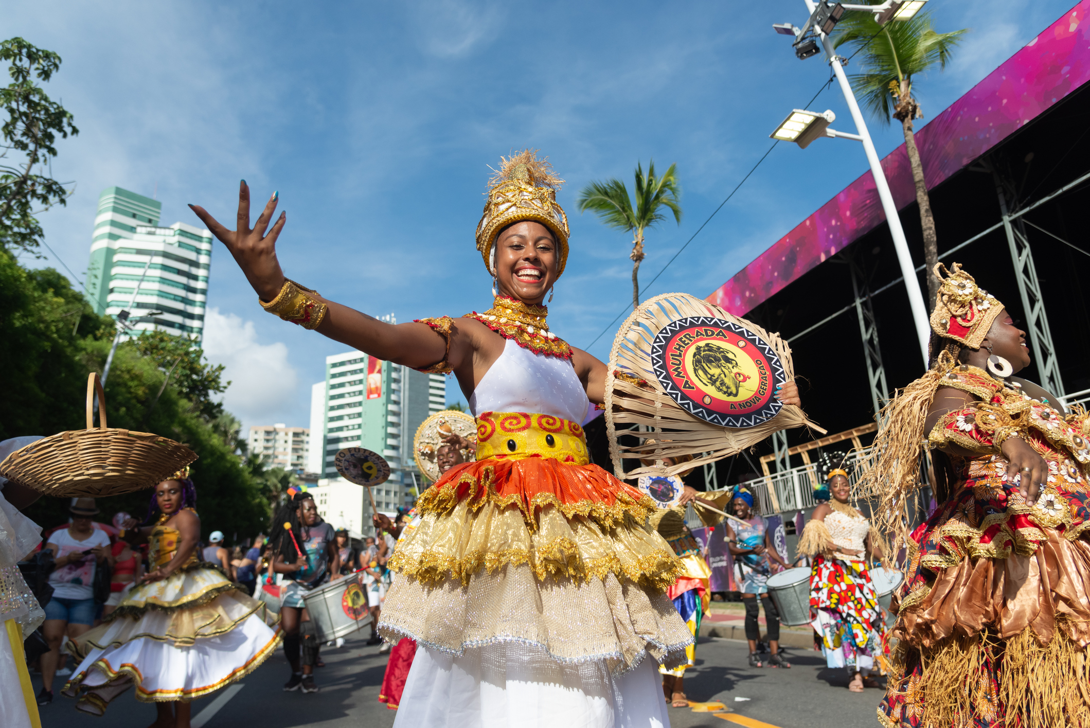 A smiling dancer in vibrant costume celebrates during a lively parade, surrounded by colorful decorations and onlookers.