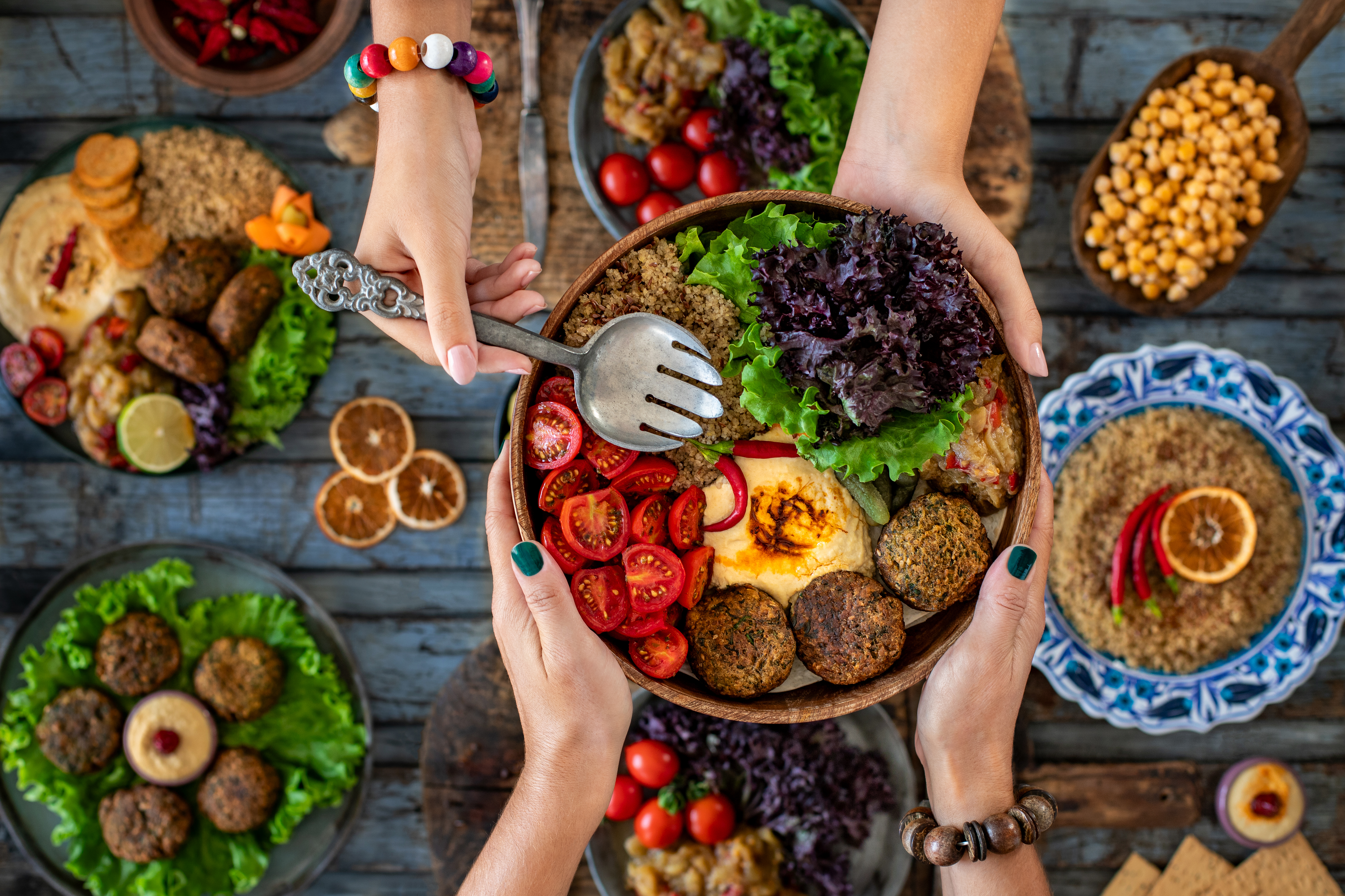 A colorful spread of salads, falafel, and vegetables, with hands reaching for a bowl on a rustic wooden table.