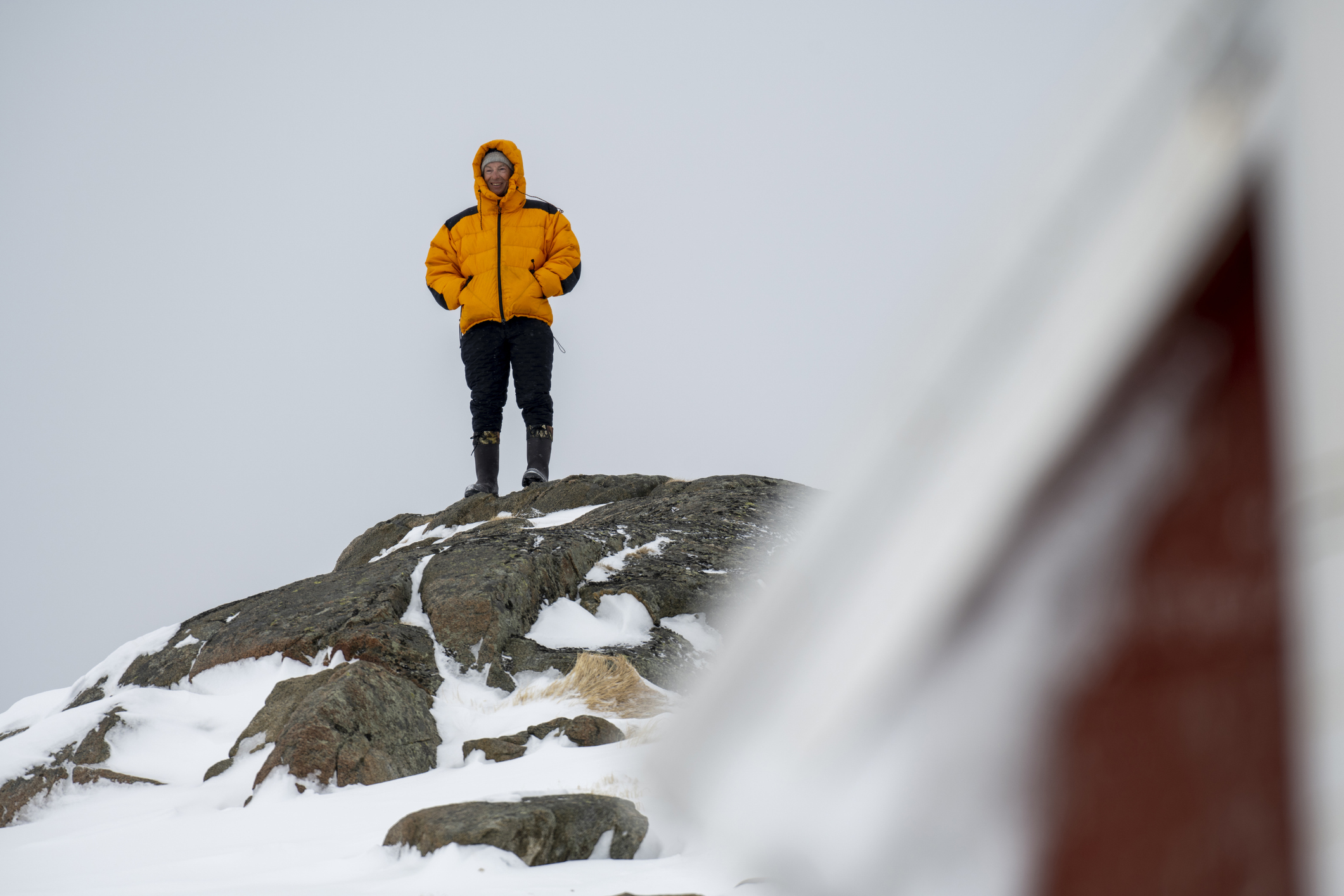 A person in a bright yellow jacket stands on a snowy rock, overlooking a winter landscape with a blurred structure nearby.