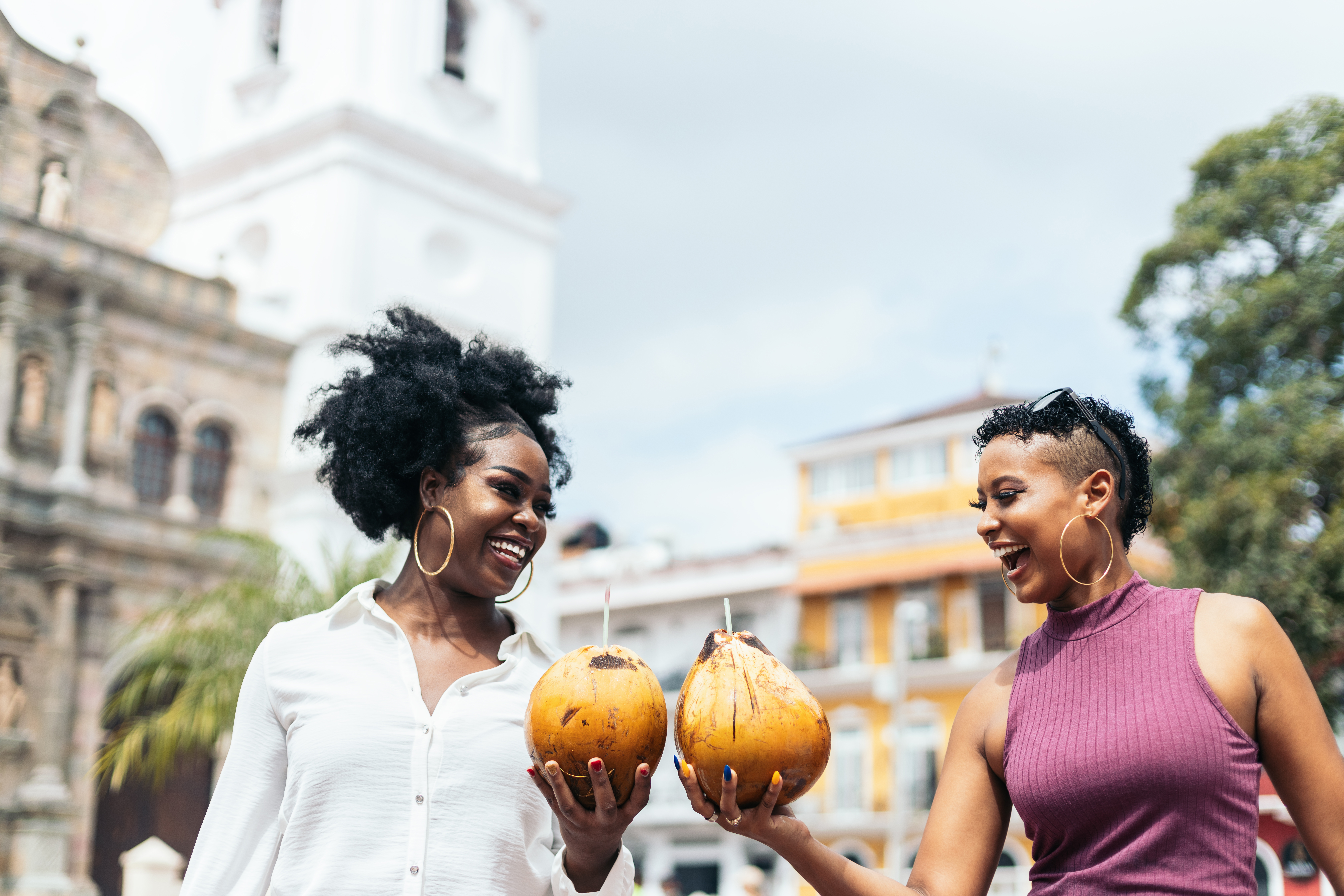 Two women smile and hold coconuts in a vibrant outdoor setting with historic buildings and greenery in the background.