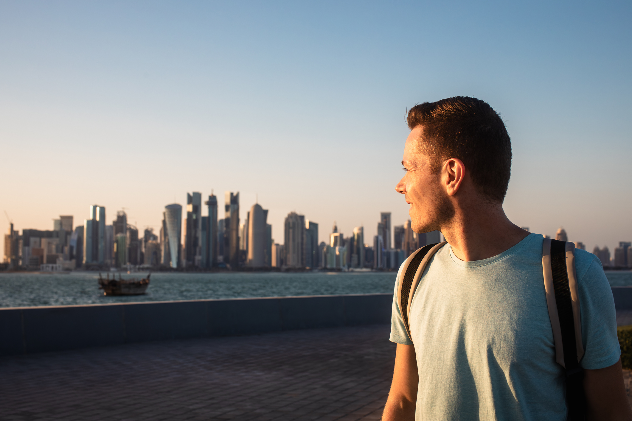 A man gazes at a city skyline during sunset, with water and a boat in the foreground.