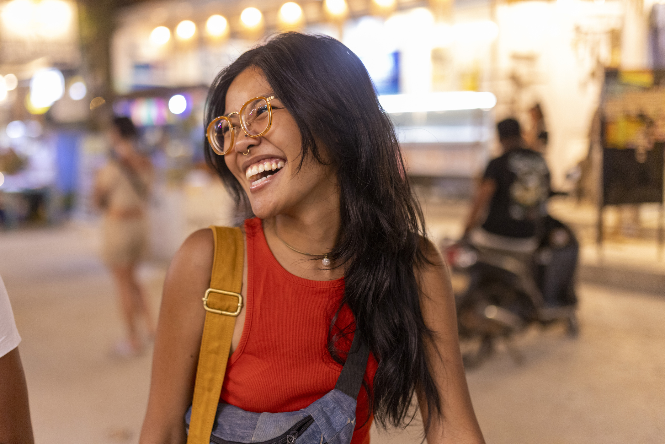 Smiling woman with long black hair, wearing glasses and a red shirt, standing in a bustling outdoor setting.