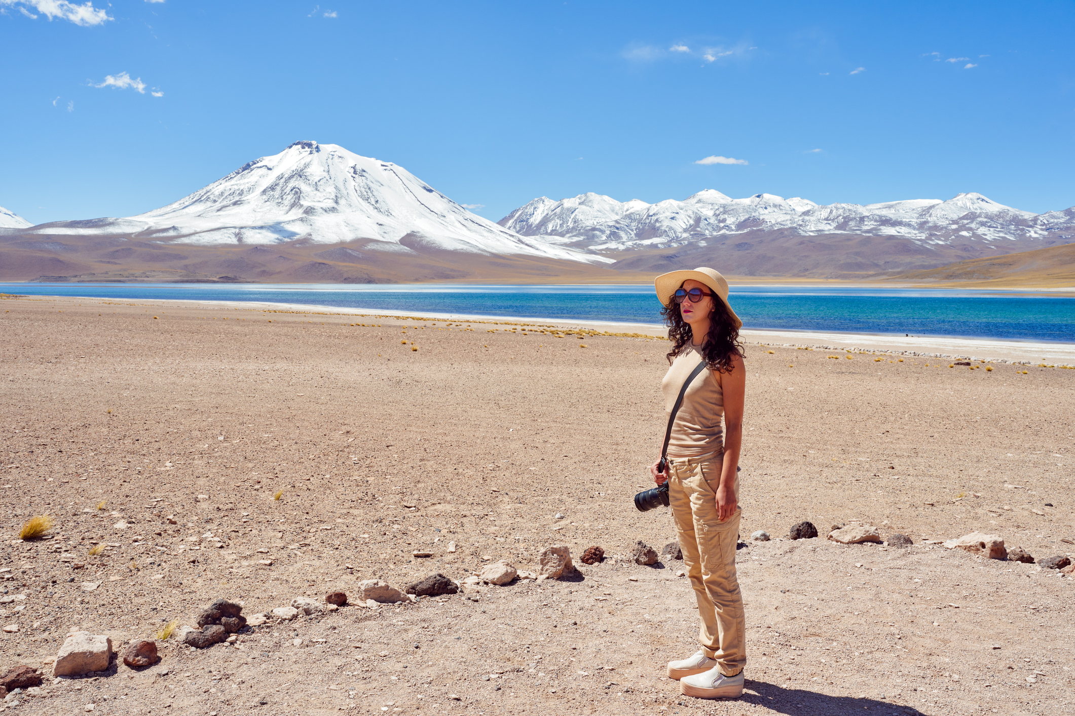 A person stands on a rocky shore by a turquoise lake, with a snow-capped mountain in the background under a clear blue sky.
