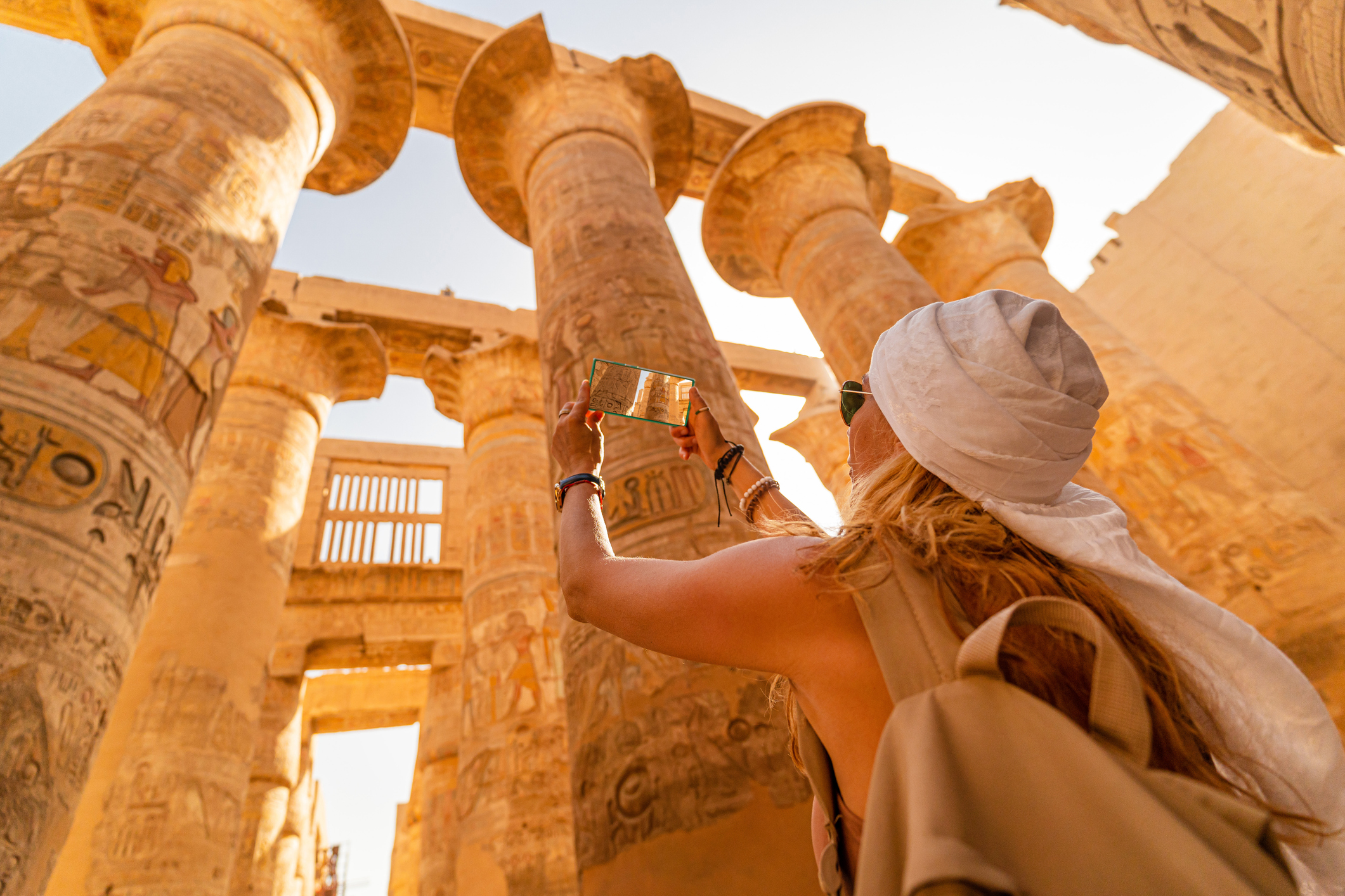 A person taking a photo of ancient columns with hieroglyphs in a sunlit archaeological site.