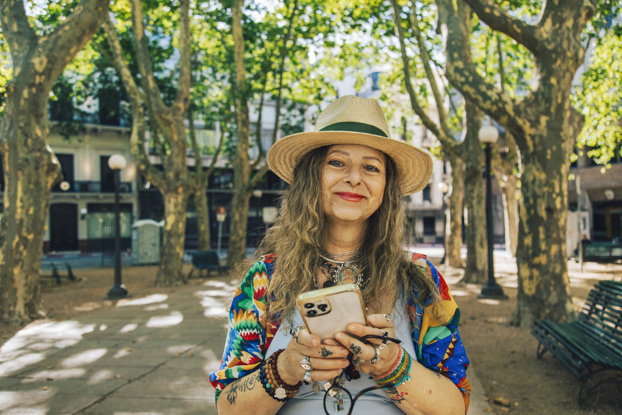 Smiling woman with long, wavy hair, wearing a straw hat and colorful clothing, holding a phone in a sunny park setting.