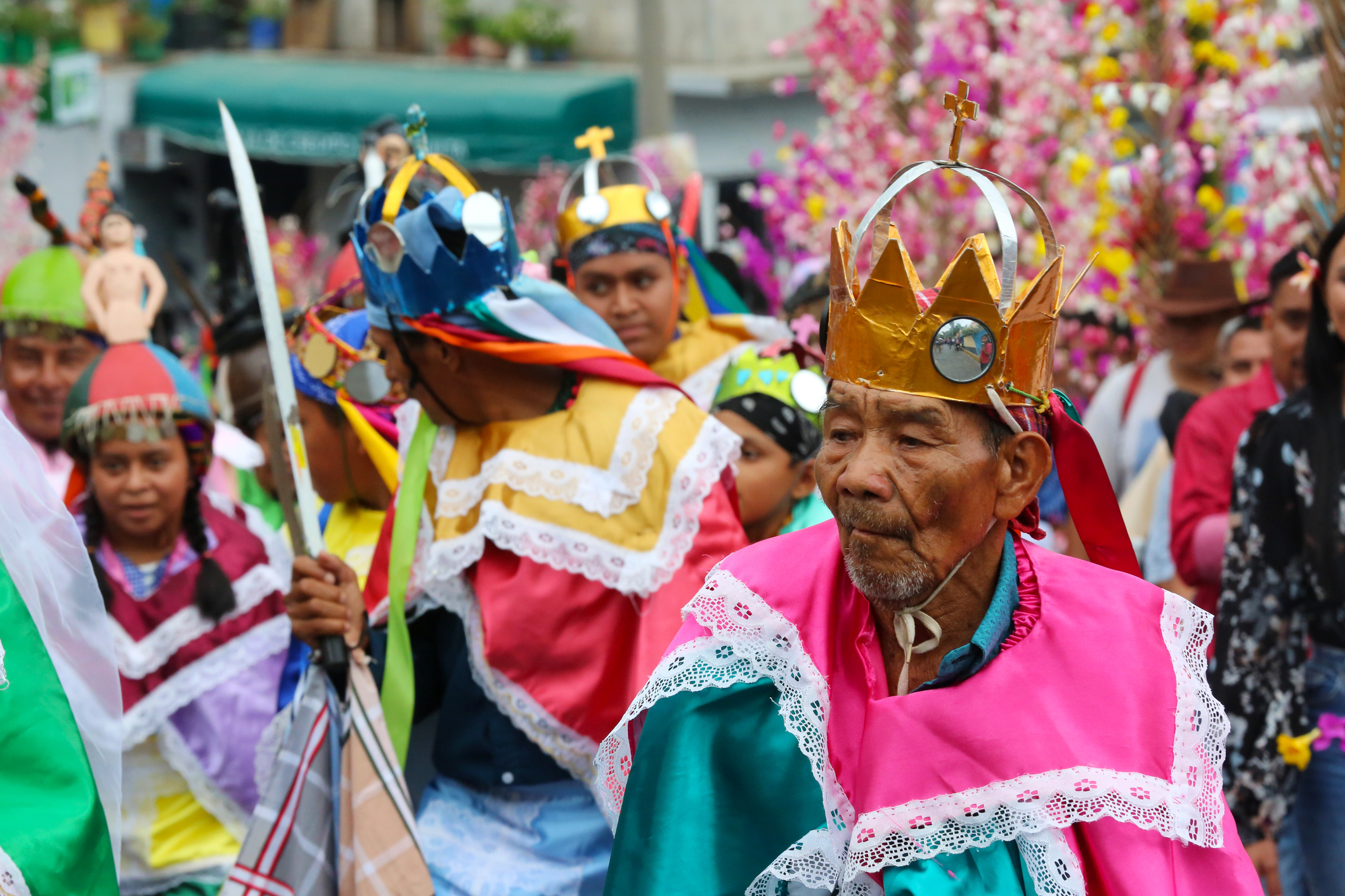 A vibrant parade featuring people in colorful costumes and crowns, celebrating with joyful expressions amidst festive decorations.