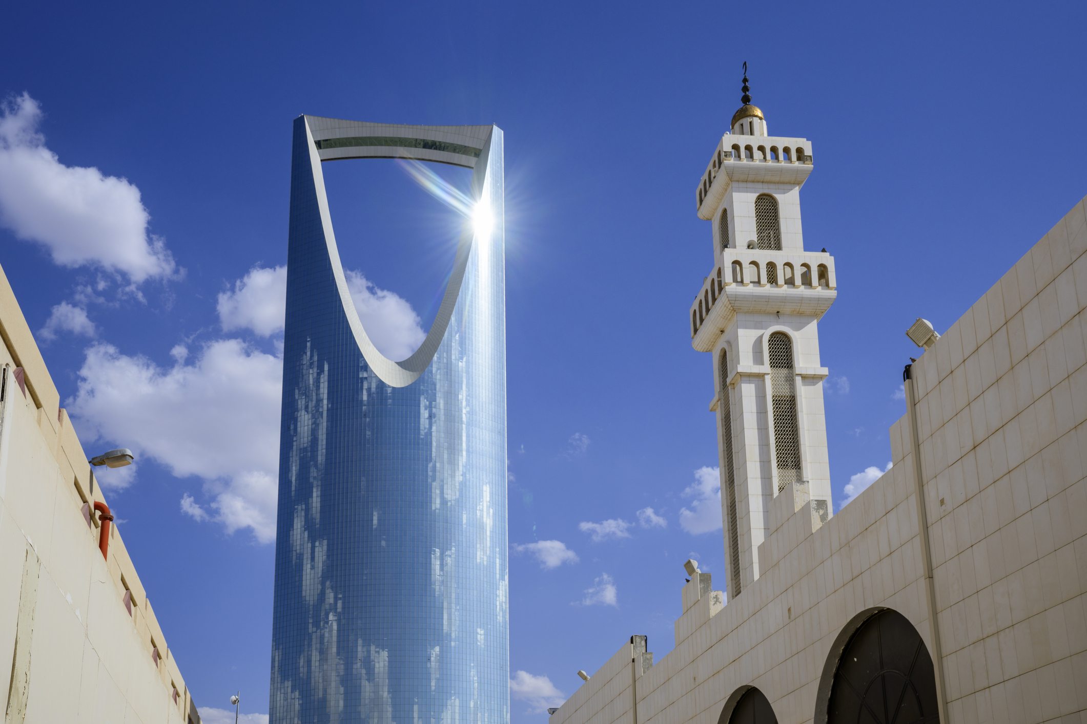 A modern skyscraper with a large opening, next to a traditional mosque tower, against a blue sky with clouds.
