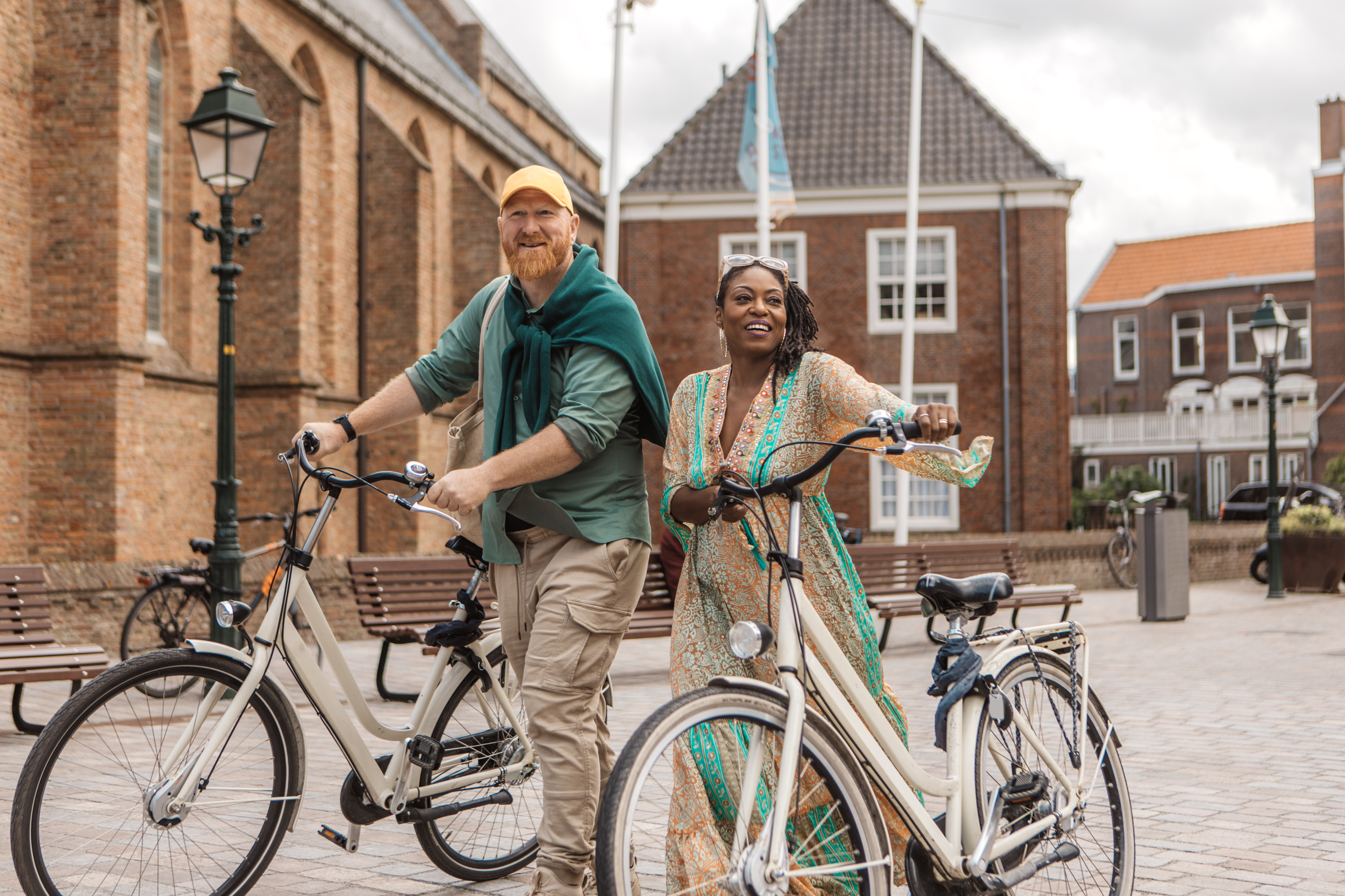 Two smiling people, a man and a woman, stand with bicycles in a charming plaza surrounded by historic buildings.