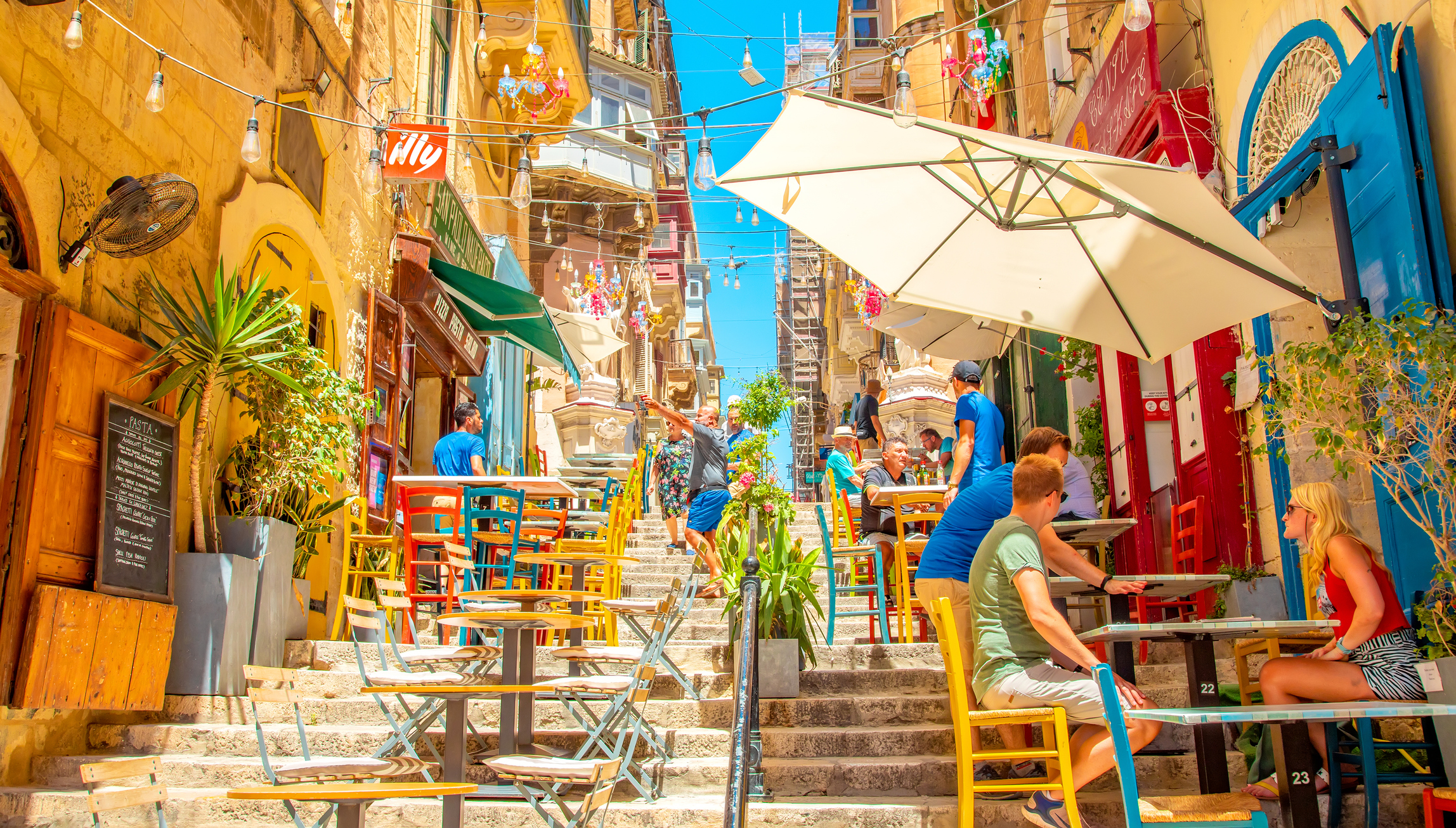 Colorful outdoor cafes line a sunlit, vibrant street, with people enjoying drinks and a bright blue sky overhead.