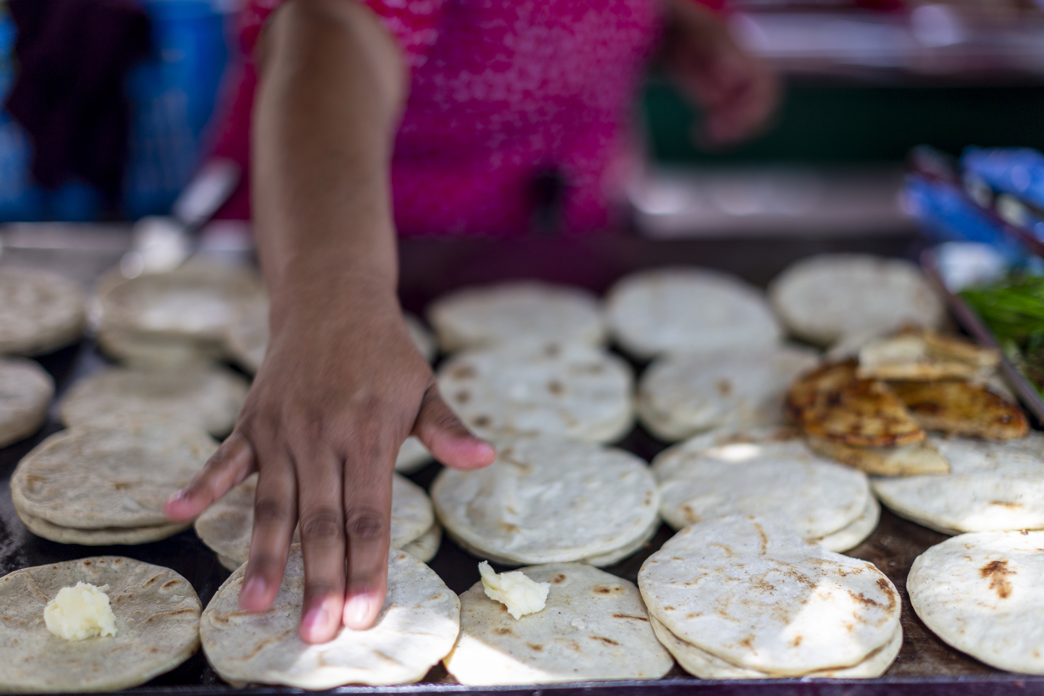 A hand spreads butter on freshly made flatbreads on a cooking surface, surrounded by more flatbreads and some grilled food.