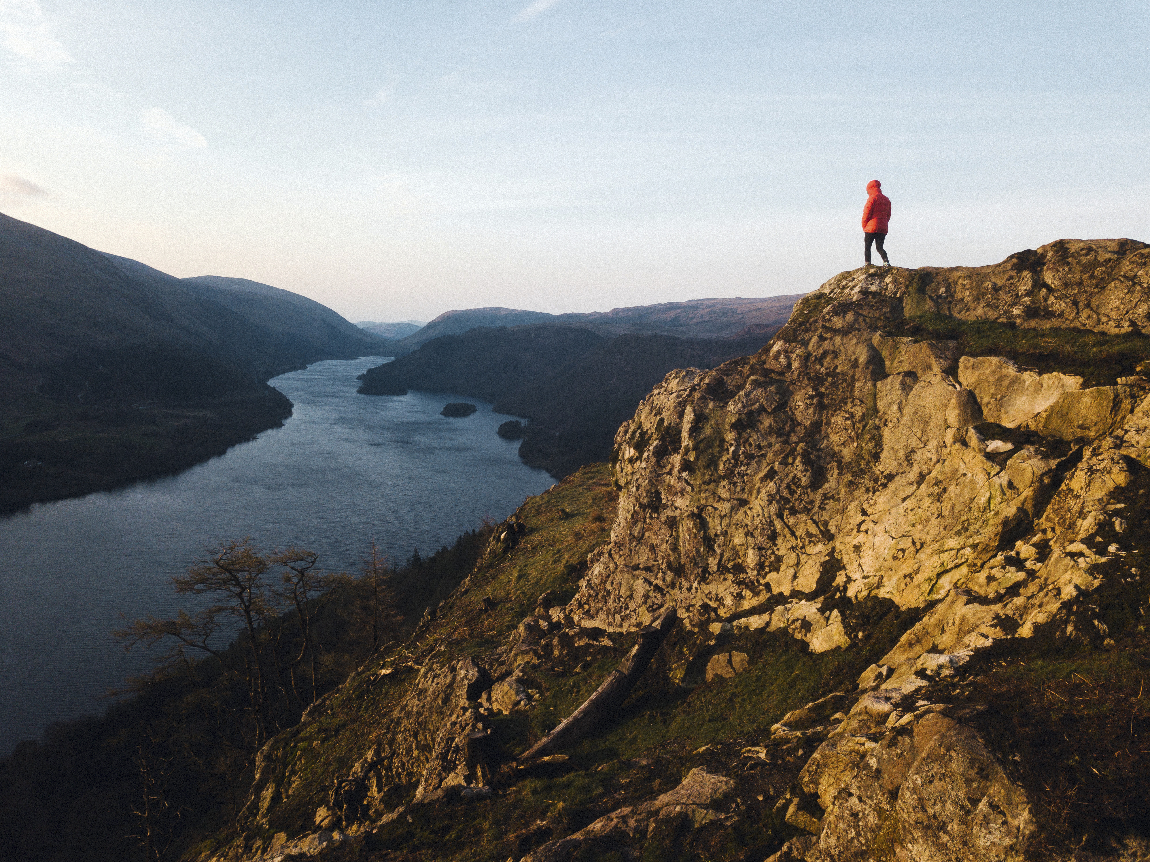 A person in a red jacket stands on a rocky outcrop overlooking a river and mountains at sunset.