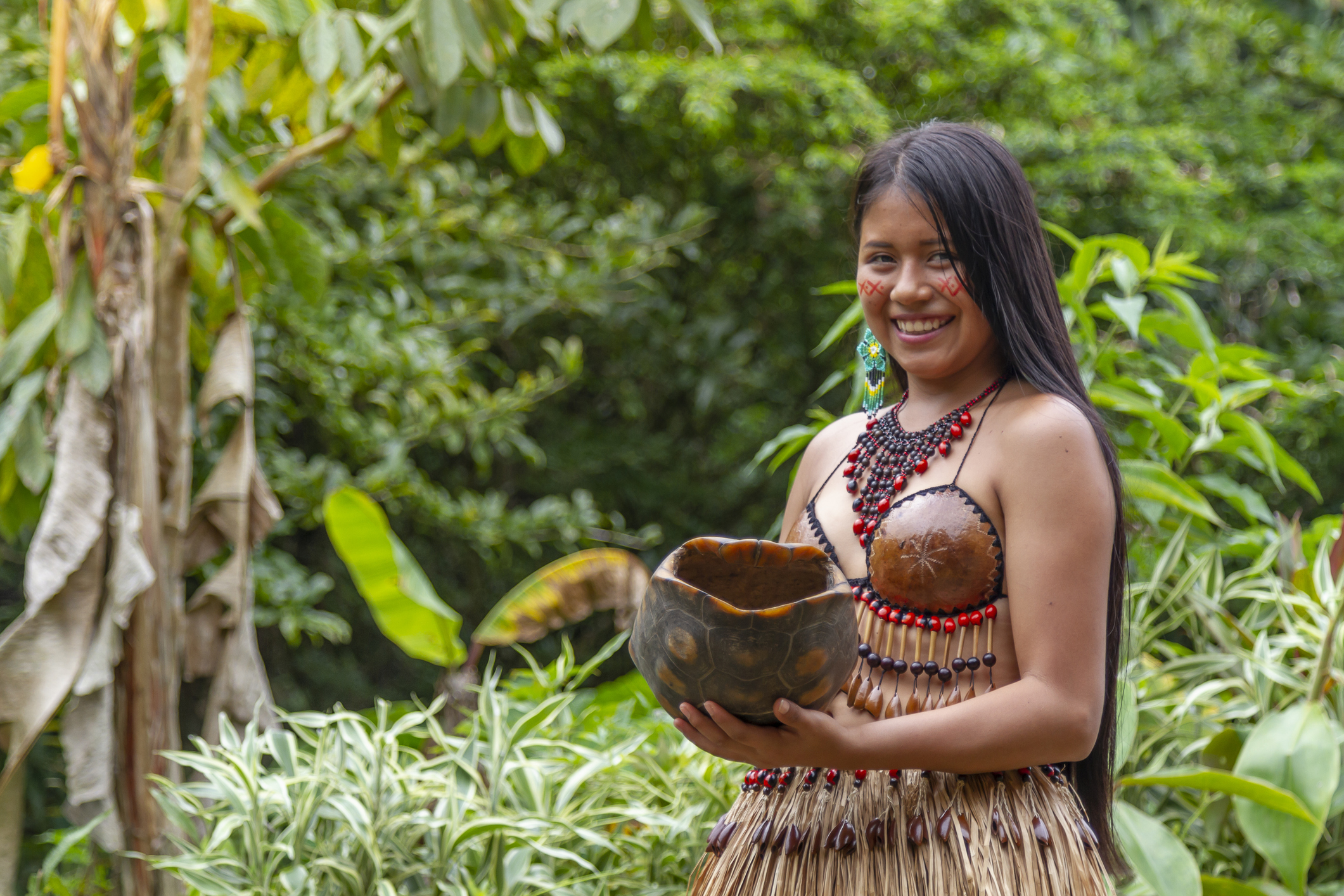 A woman in traditional attire smiles while holding a decorative bowl surrounded by lush greenery.