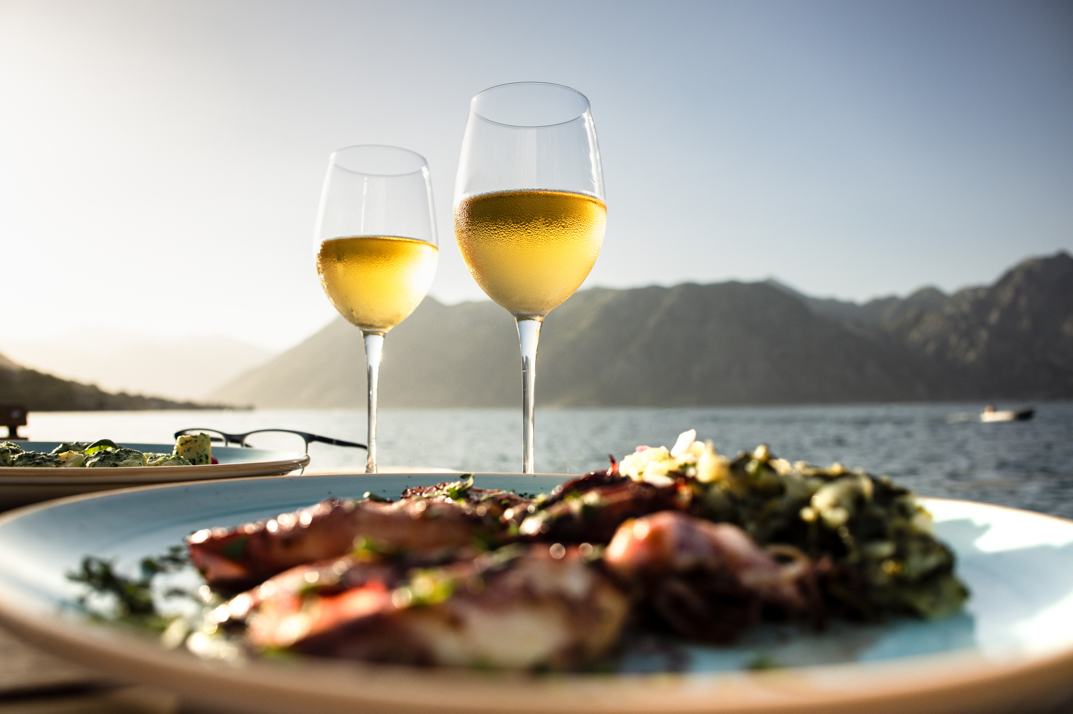 Two glasses of white wine in front of a scenic view with mountains, alongside a plate of seafood and greens.