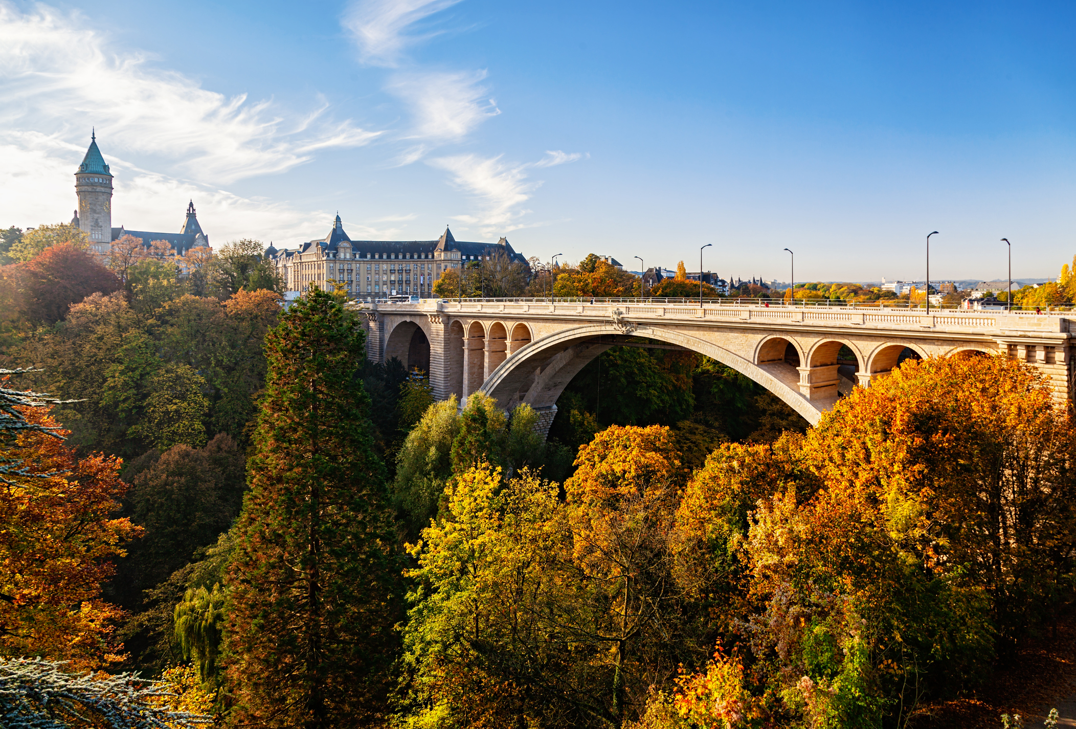 Scenic view of an arched bridge surrounded by colorful autumn trees and historic buildings against a blue sky.