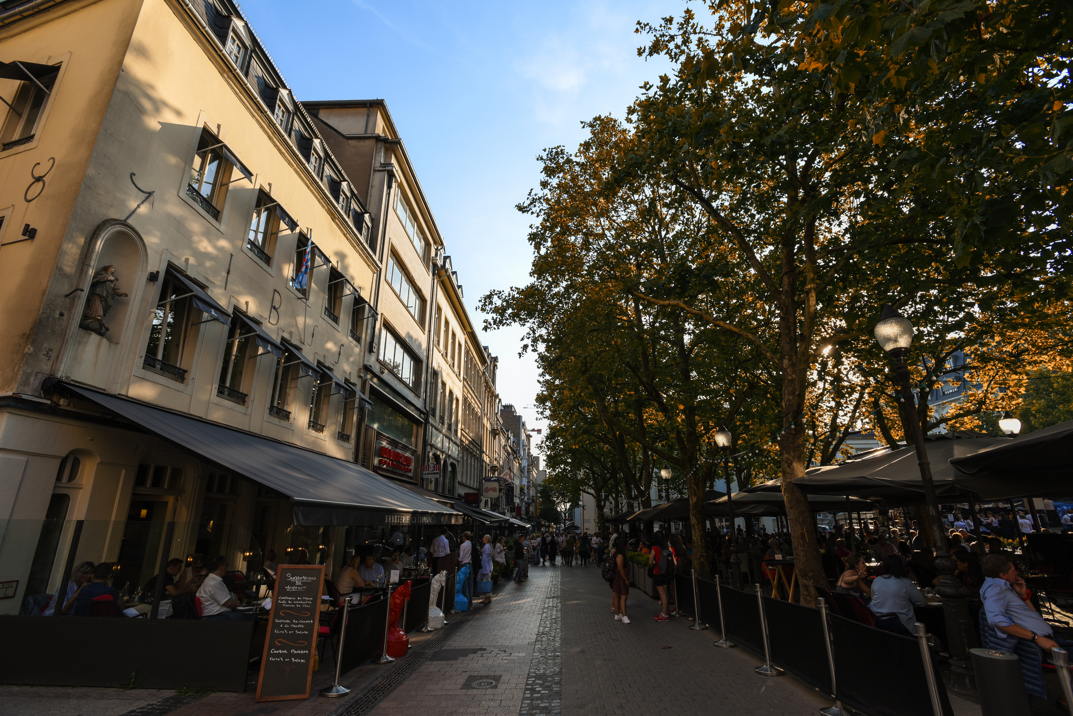 Street lined with cafes and trees, busy with people enjoying dining and conversation in a warm, autumn afternoon.