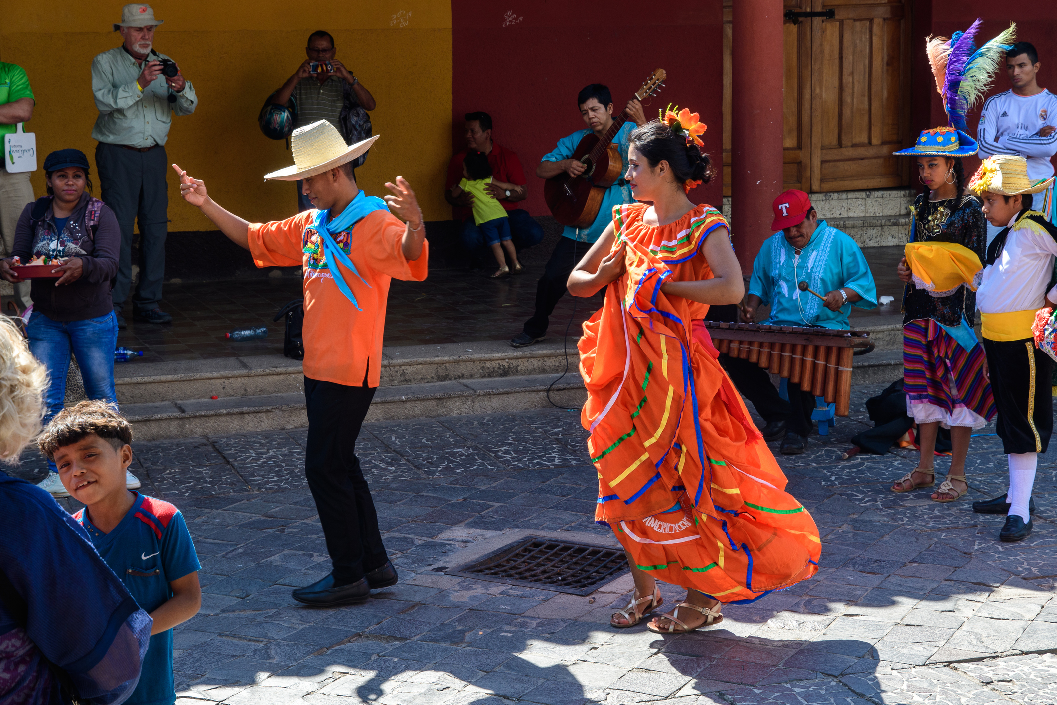 Colorful dancers perform a traditional dance in a lively outdoor setting, with musicians and a crowd around them.