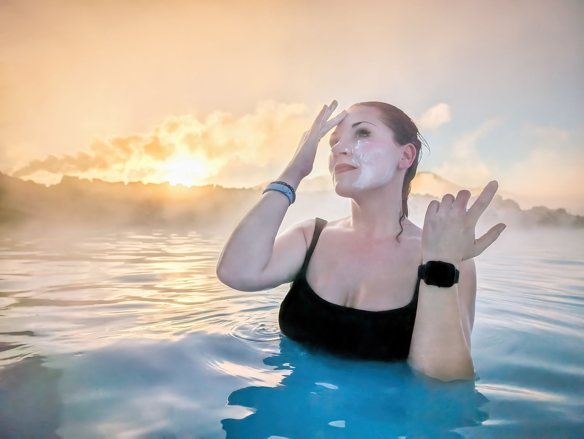 A person in a black swimsuit applies facial cream while standing in steam-filled water at sunset.