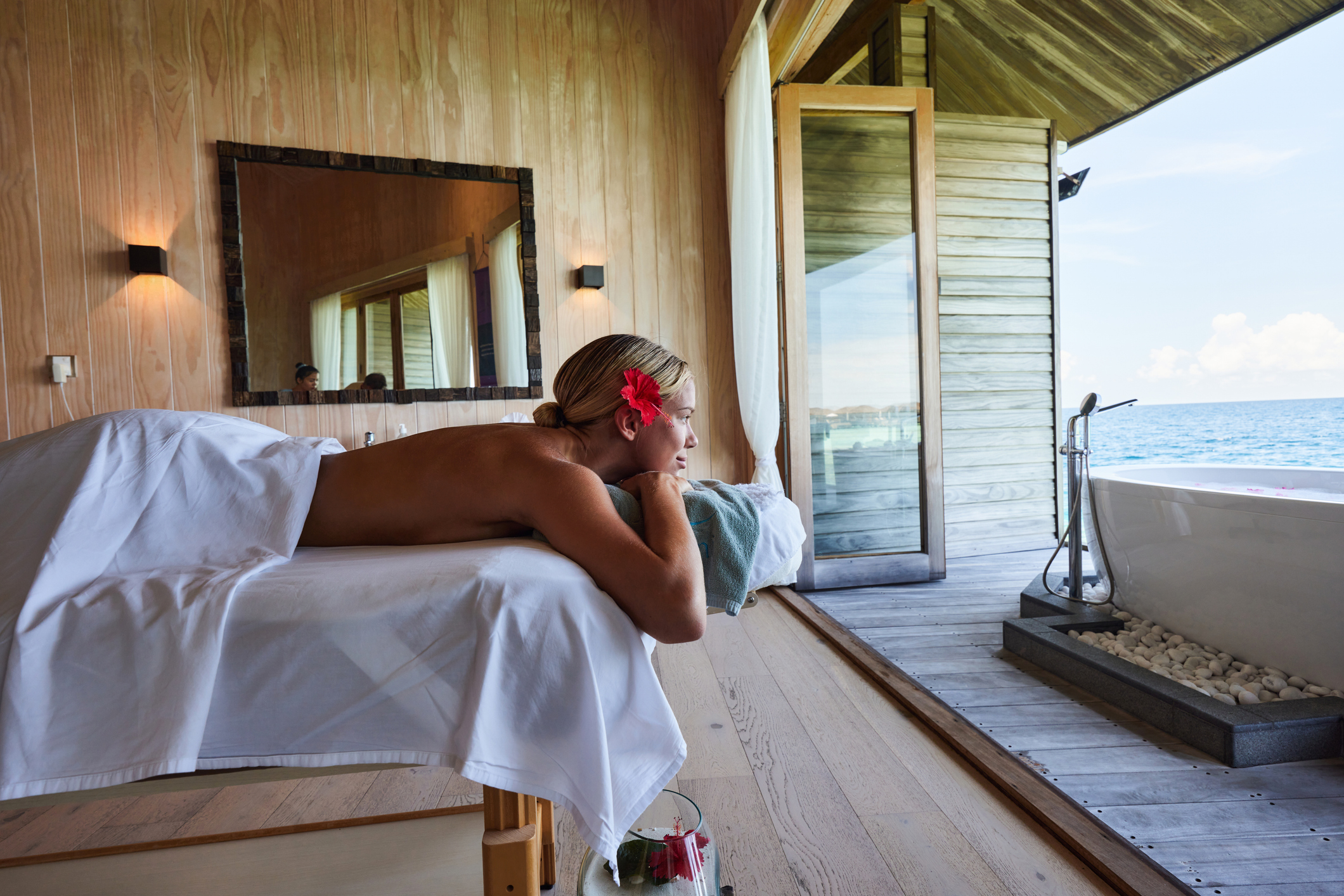 A person enjoys a relaxing massage in a serene spa with a view of the ocean, adorned with a flower in their hair.