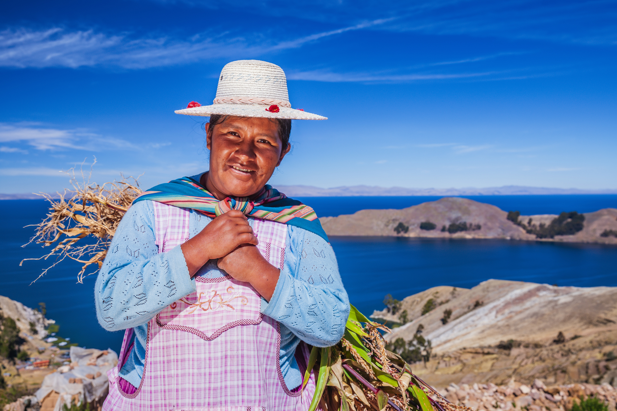 A smiling woman in traditional attire stands near a lake, holding plants, with mountains and a clear blue sky in the background.