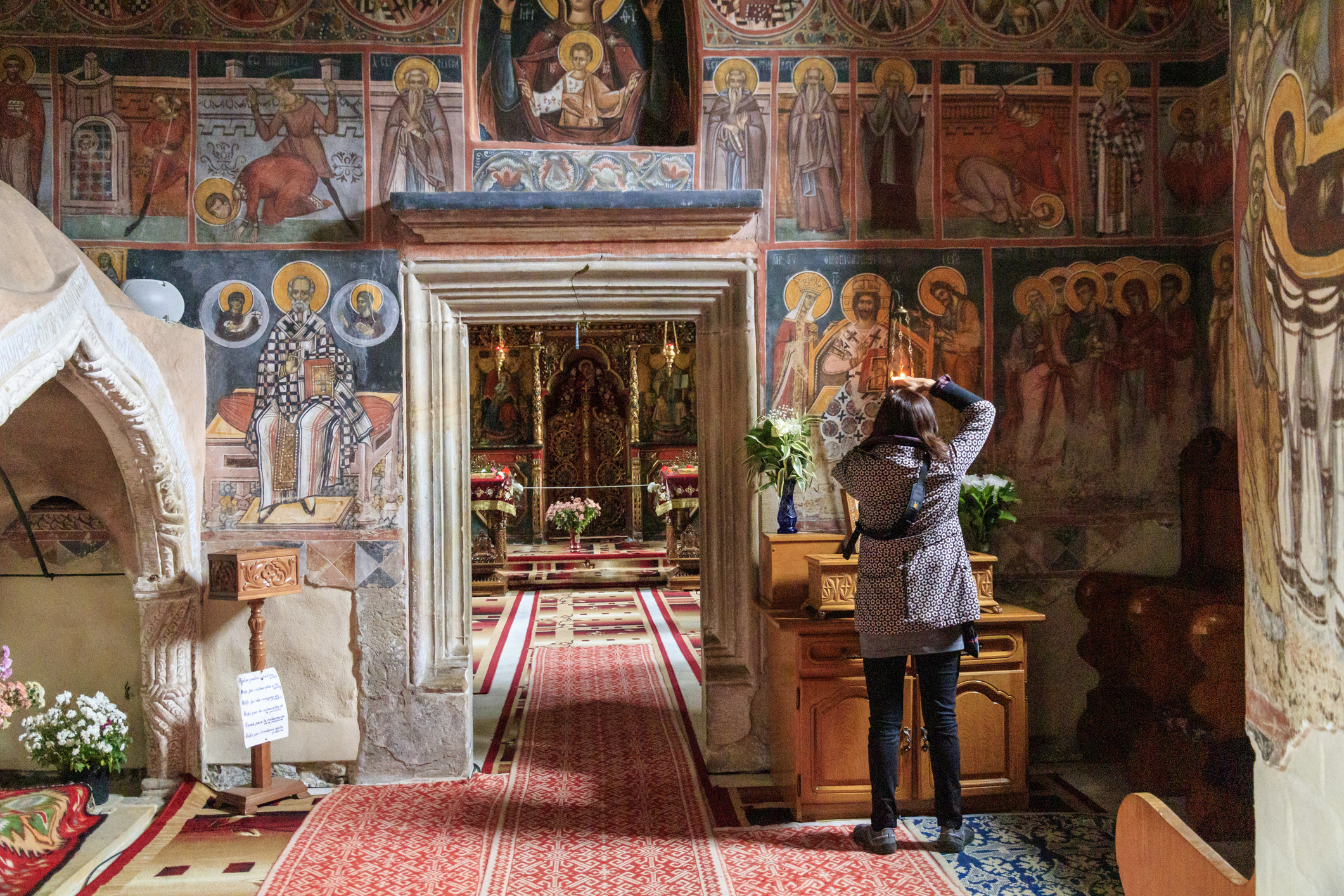 A person admires colorful frescoes inside a church, with floral arrangements and a dimly lit altar in view.