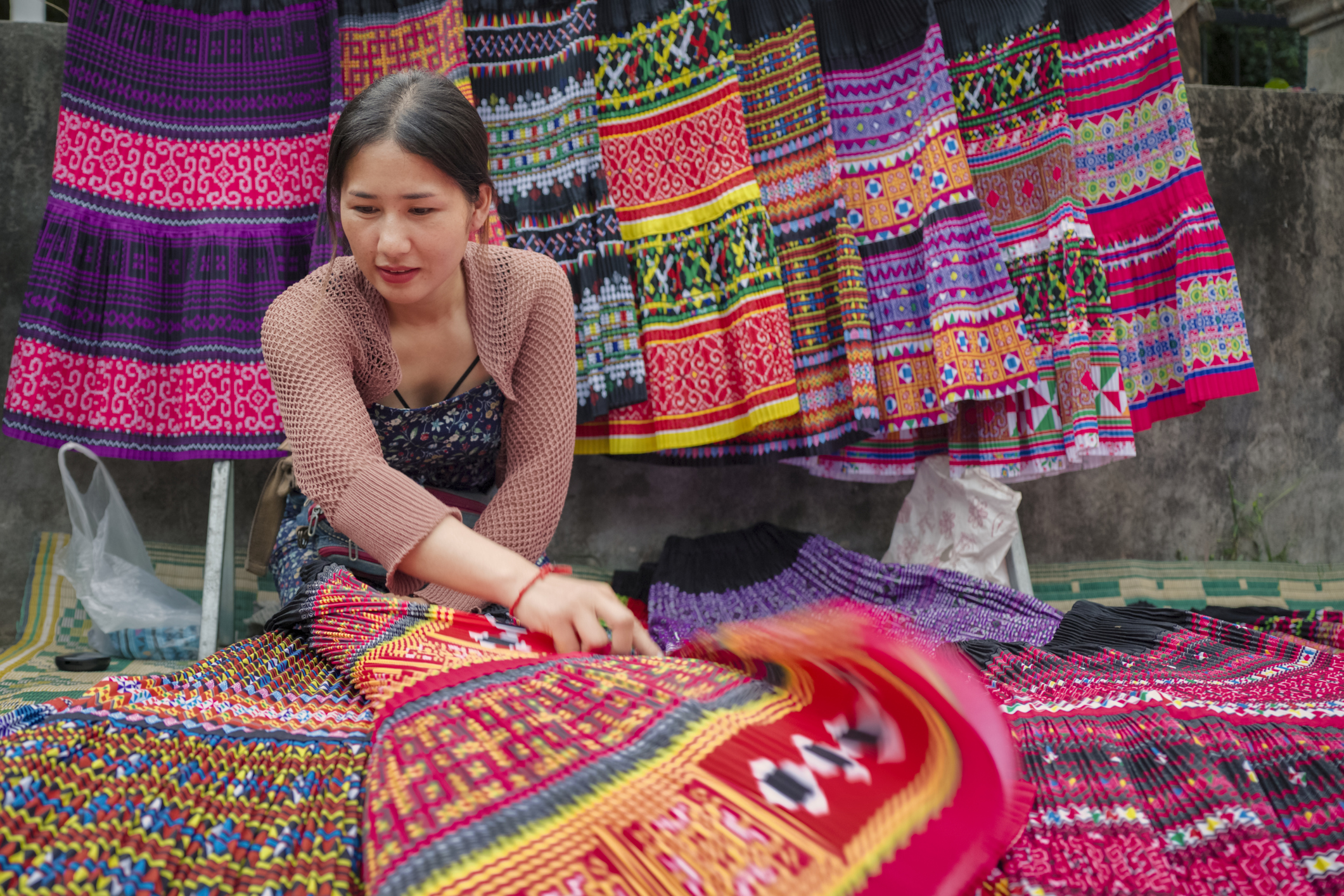 A woman arranges colorful, patterned textiles at a market, with vibrant traditional garments hanging in the background.