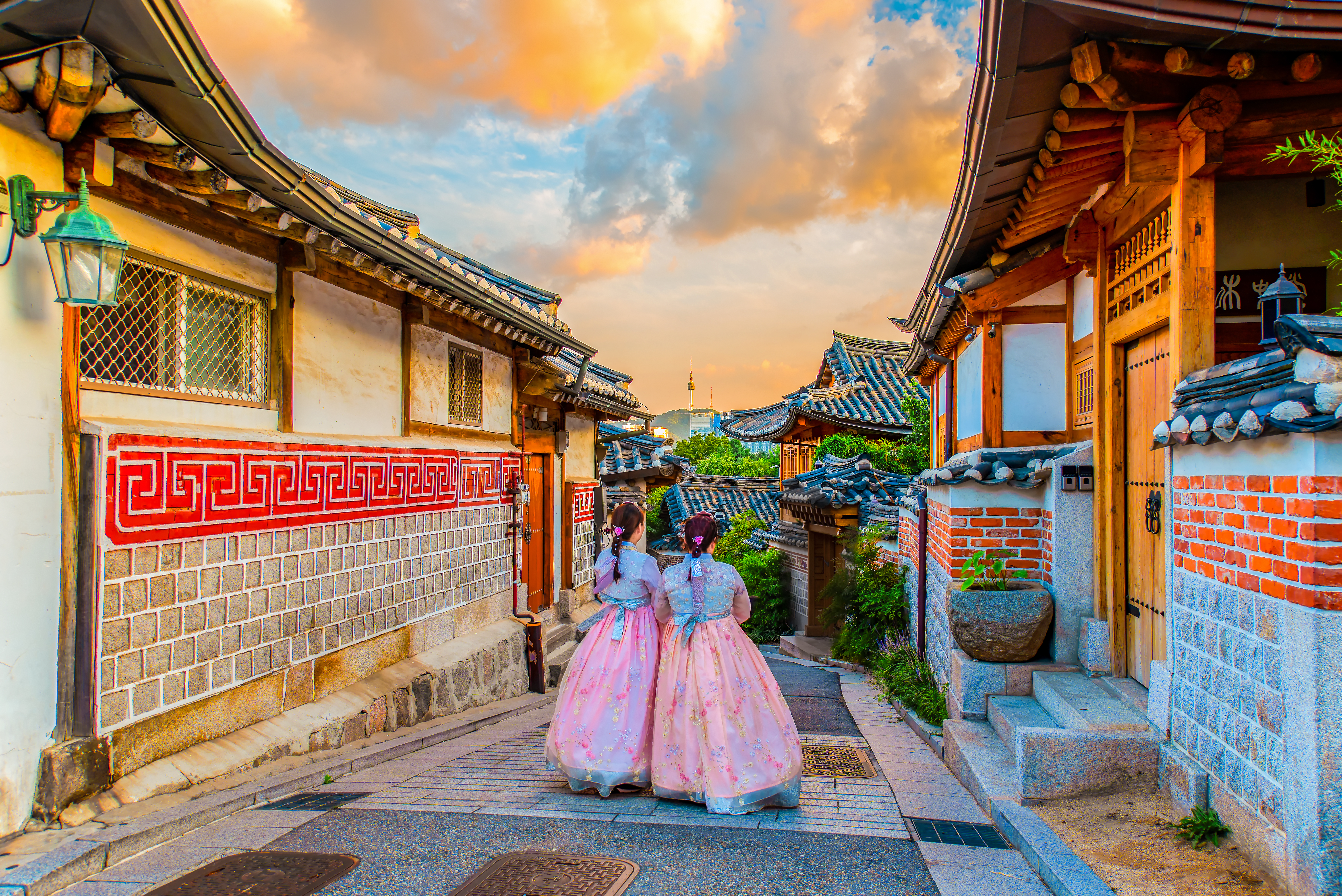 Two girls in traditional attire stroll through a picturesque alley lined with wooden houses and colorful sky at sunset.
