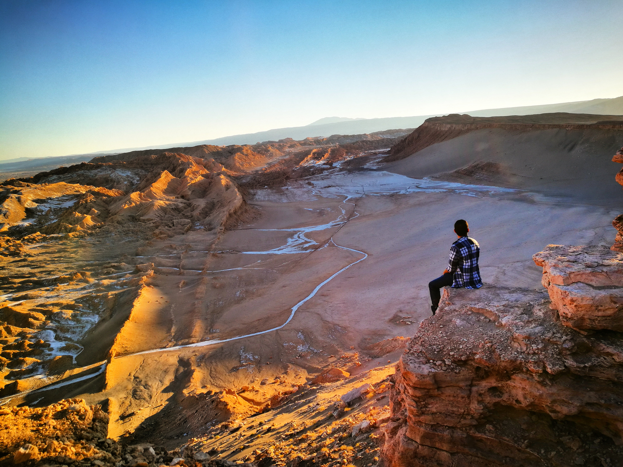 A person sits on a rocky ledge, overlooking a vast desert landscape at sunset.