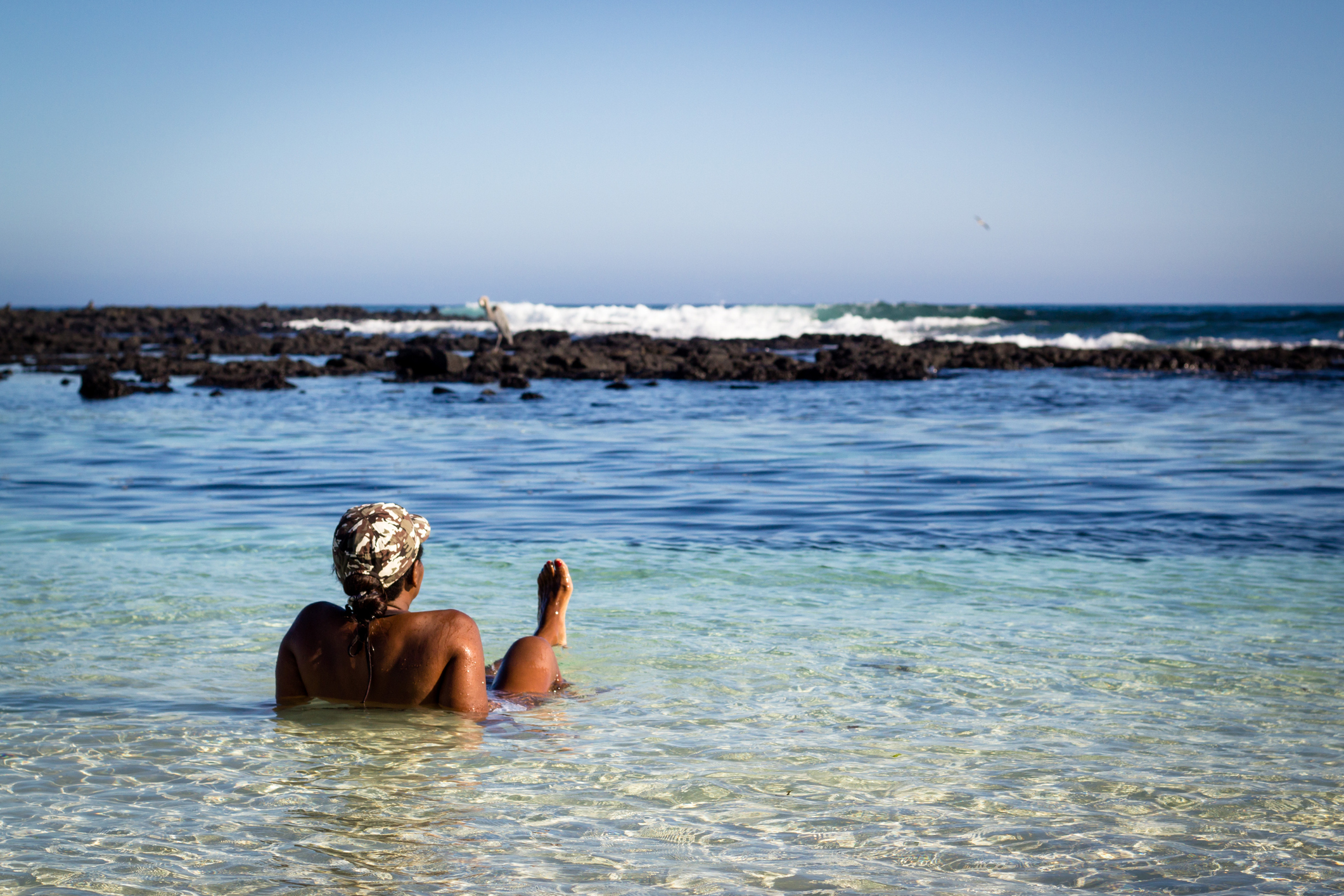 A person relaxes in shallow water at a beach, facing the ocean and surrounded by rocky shoreline under a clear sky.