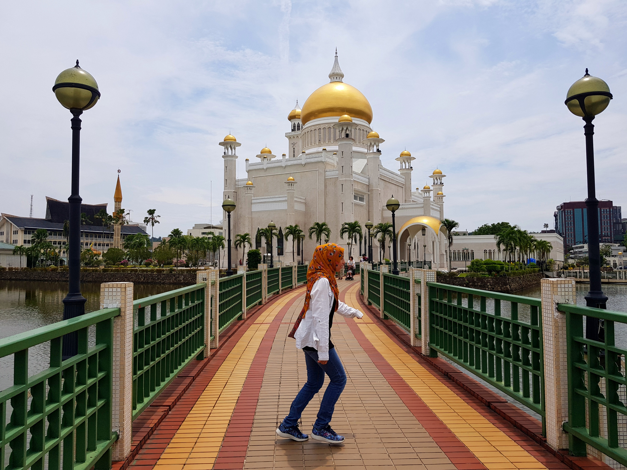 A person with orange hair walks on a bridge toward a grand mosque with golden domes, surrounded by water.