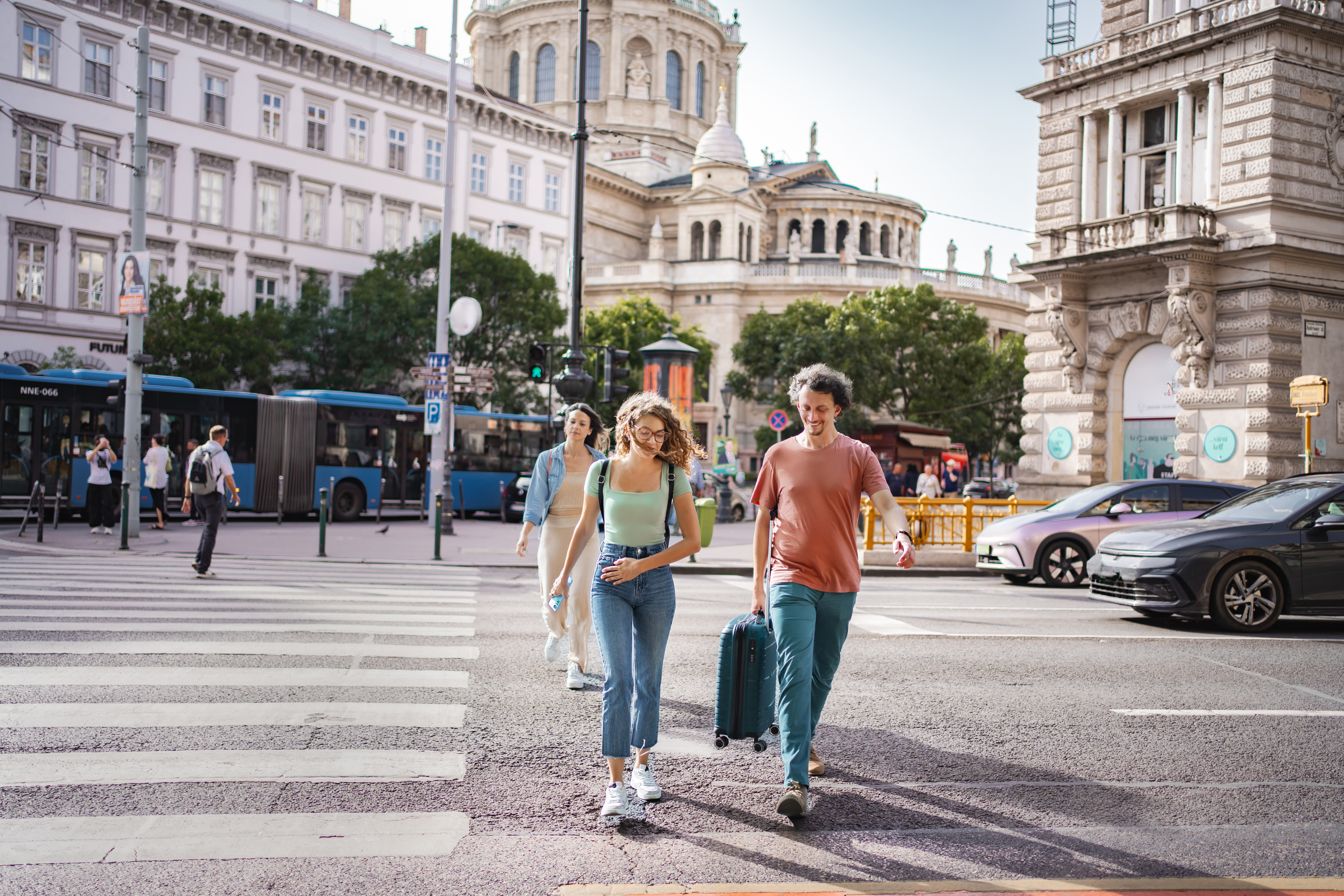 A group of three people walk across a street with a suitcase, surrounded by city buildings and buses.
