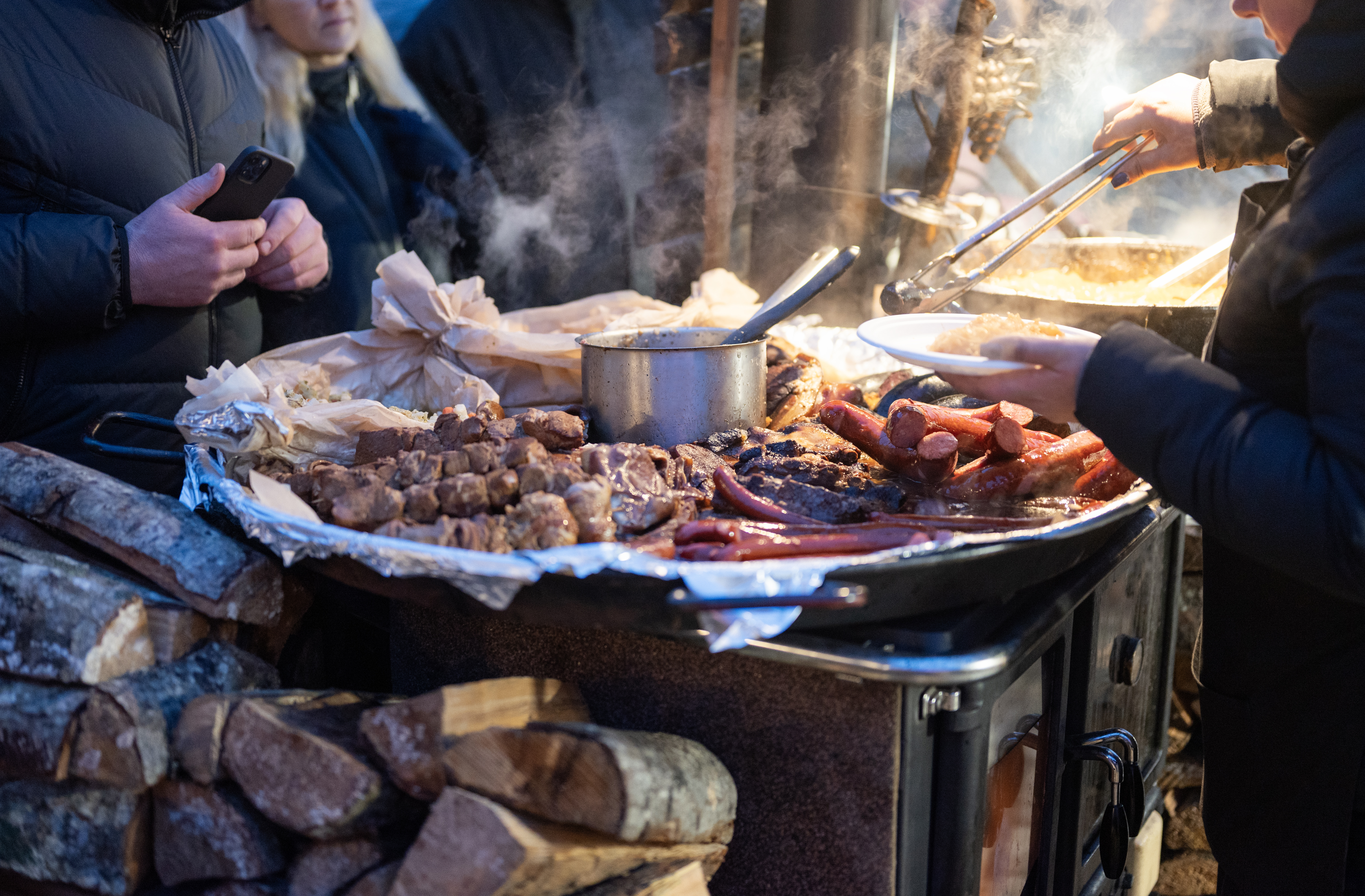 Close-up of a food stall with various meats being served, steam rising, and people in the background.
