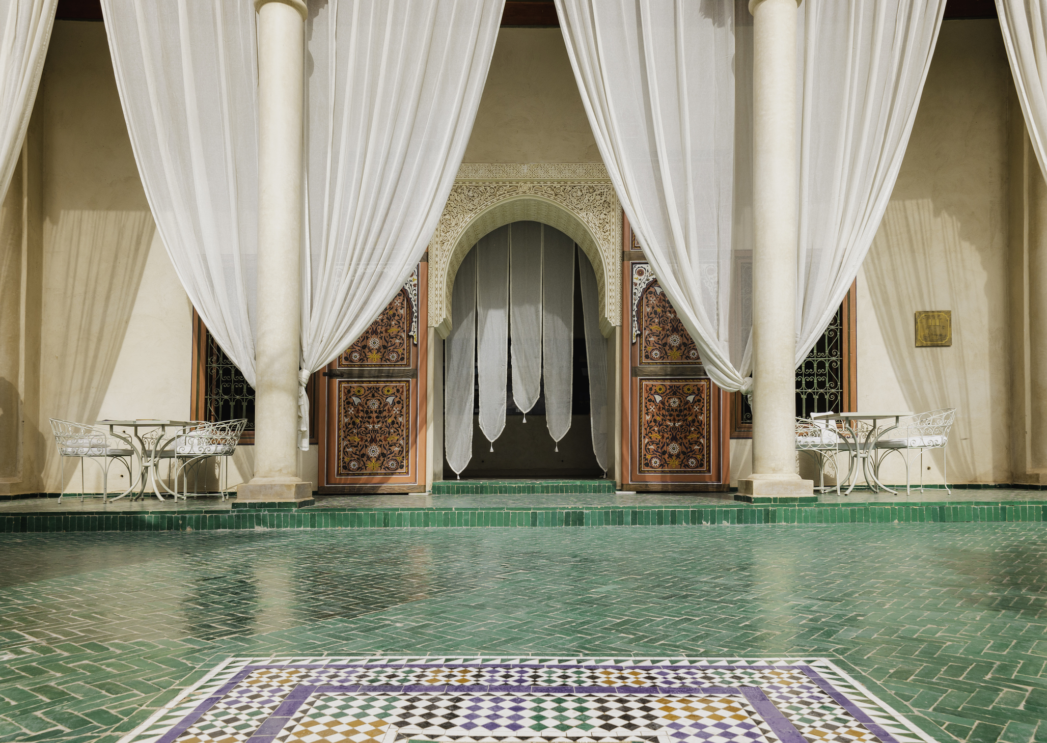 Elegant interior with green tiled floor, sheer drapes, and intricately carved wooden doors in a Moroccan-style setting.