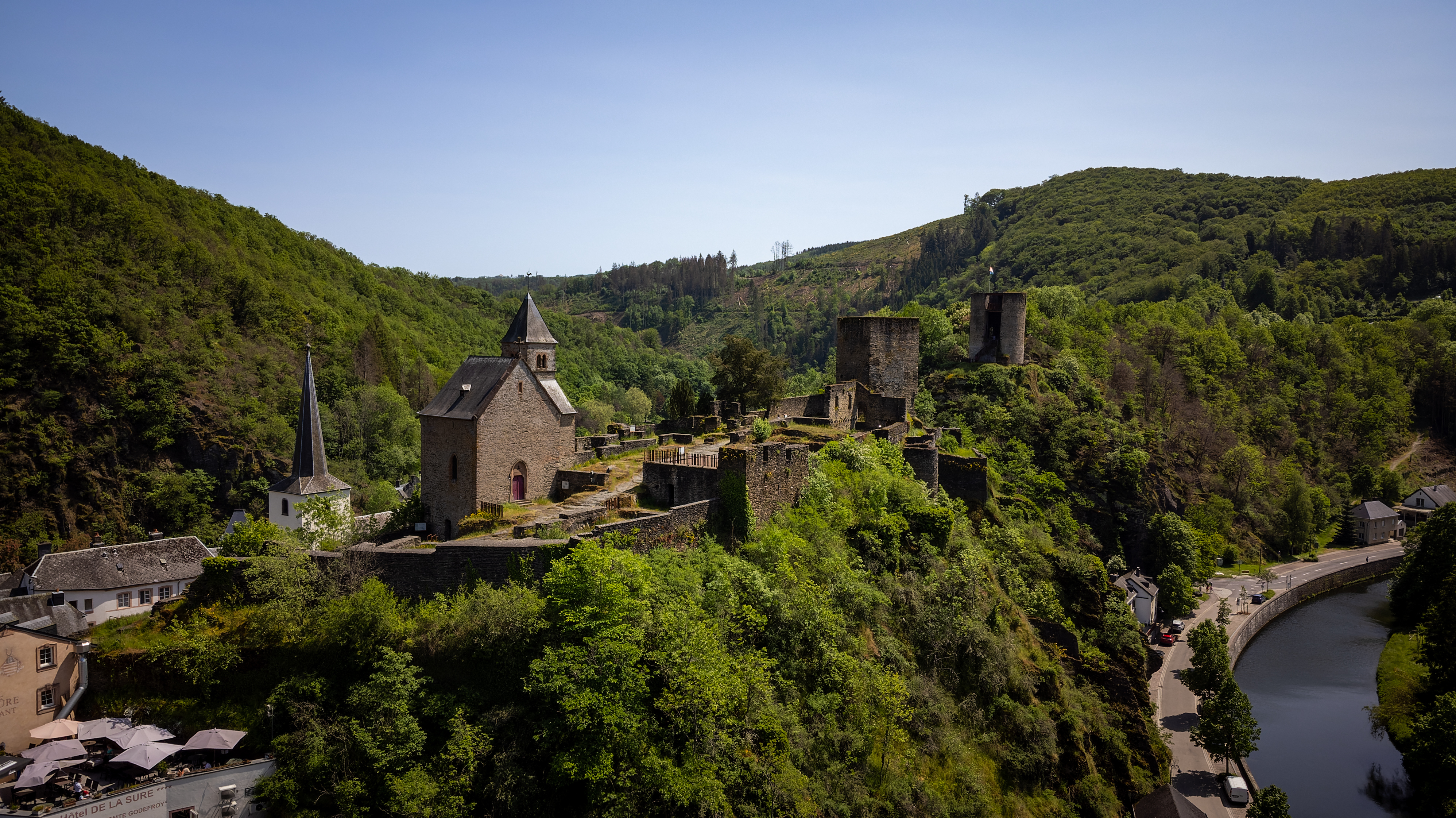 A medieval castle on a hill surrounded by lush greenery, overlooking a winding river and nearby village.